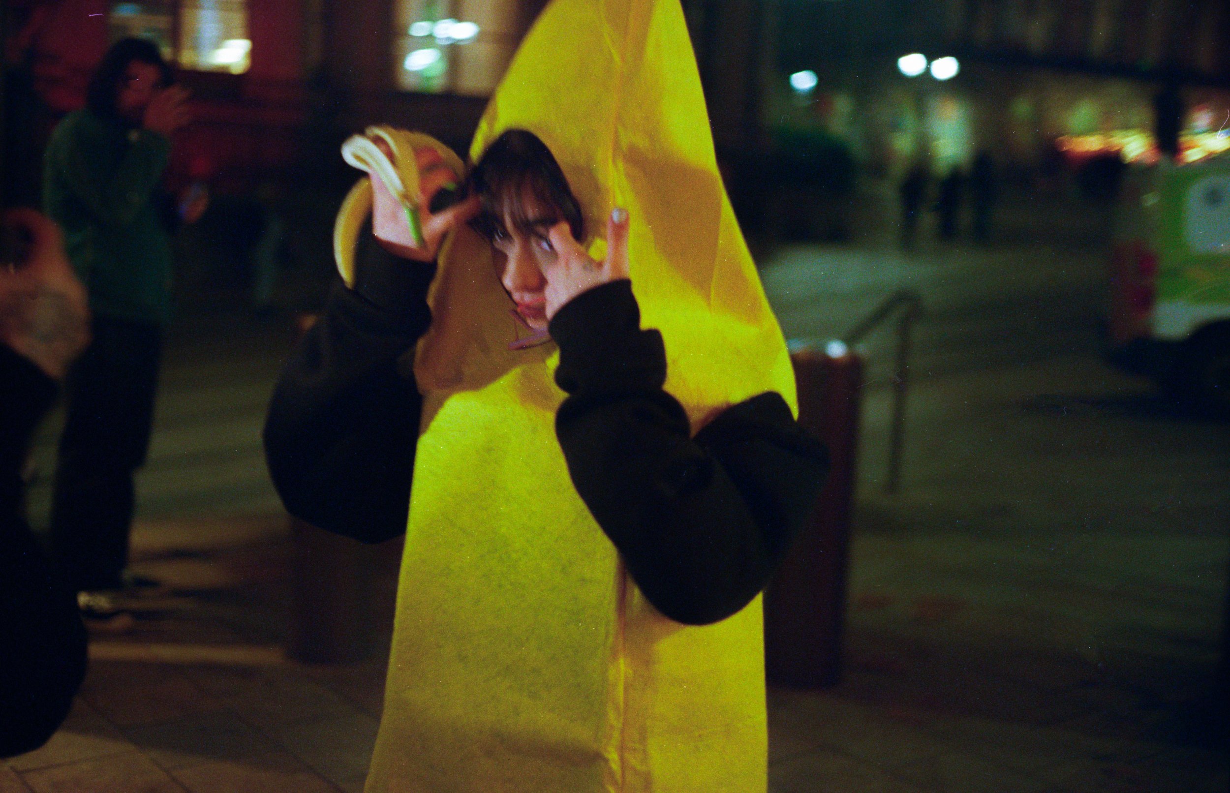 Person dressed in a banana costume making a peace sign with their hand at night on a city street.