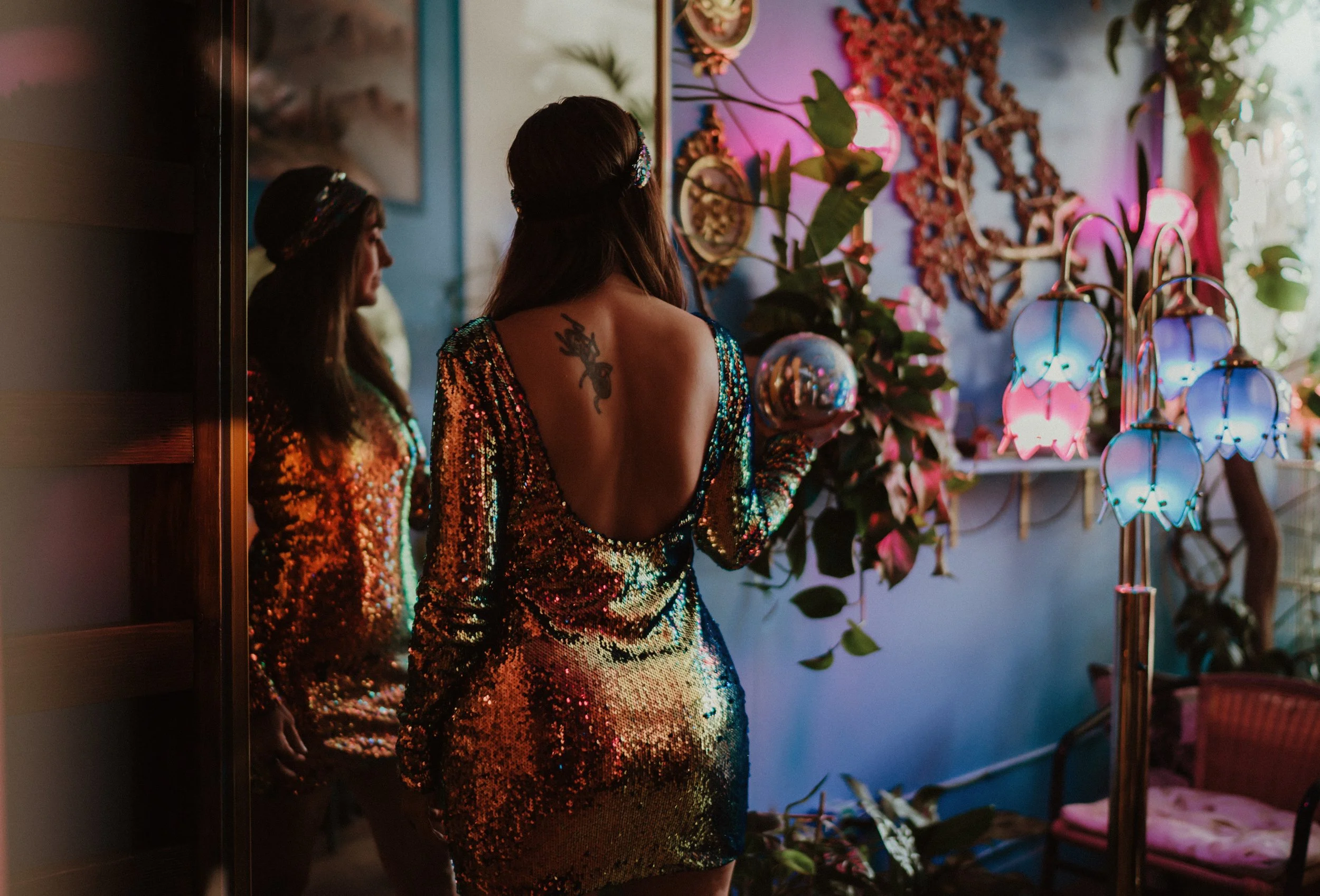 A woman in a sequined dress standing in a room with colorful lighting, plants, and decorative lamps, seen from behind.