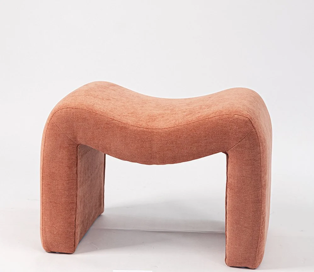 A modern upholstered stool with a curved, wave-like shape in a dusty pink fabric, set against a white background.