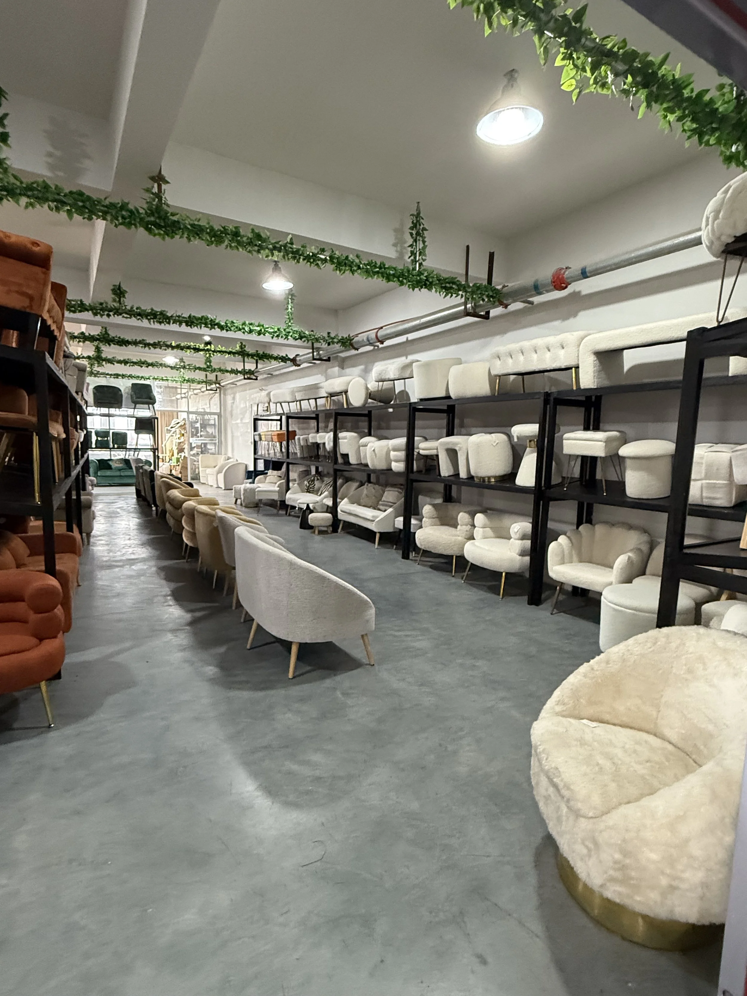 Showroom with various chairs and sofas on shelves and on the floor, with ceiling green garlands and industrial lighting.