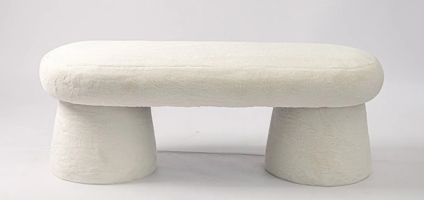 A small, white, upholstered bench with rounded top and two thick cylindrical legs.