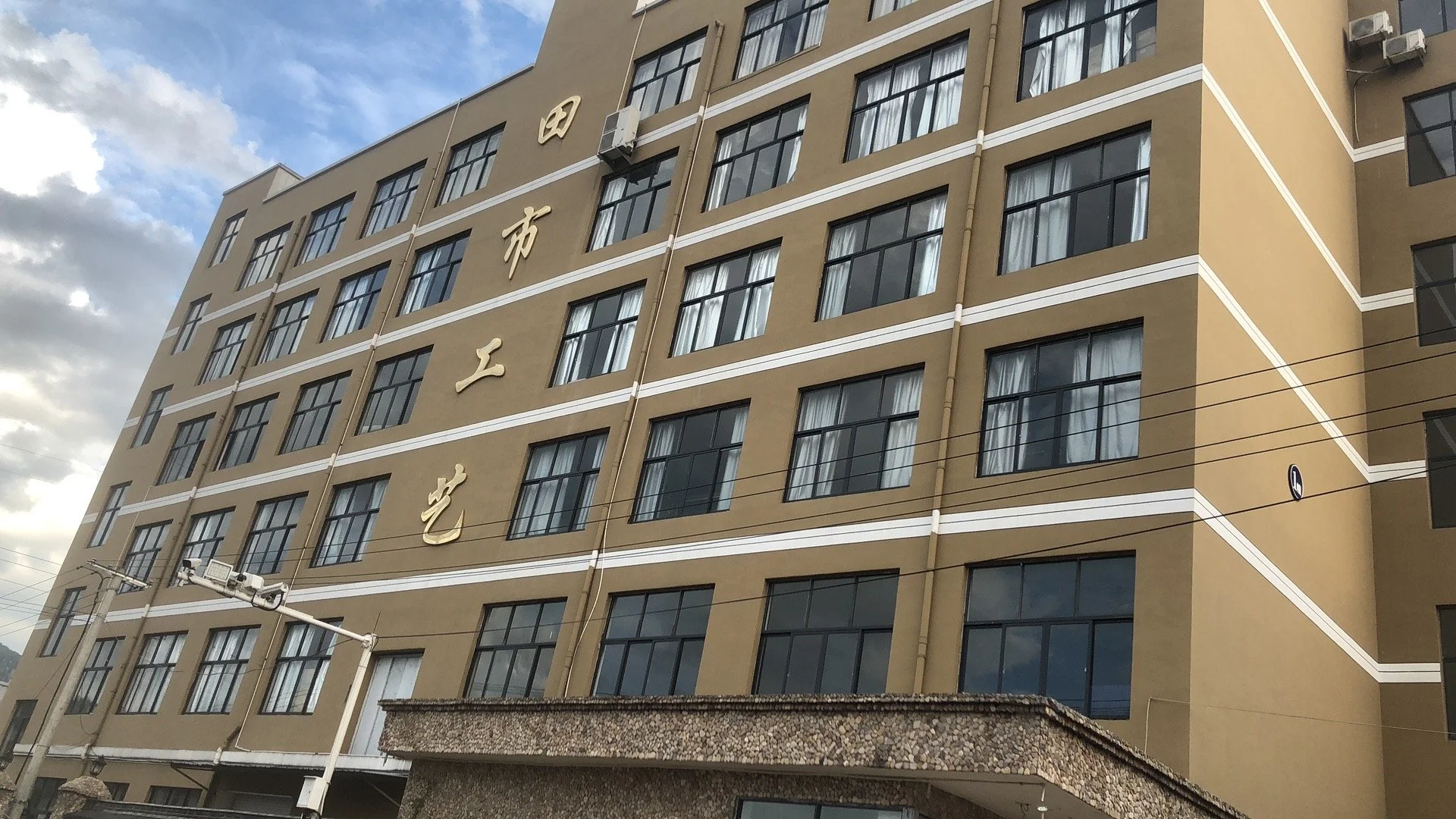 A multi-story beige building with black window frames, white trim, and Chinese characters on the side. There are power lines in front of the building and a partly cloudy sky overhead.