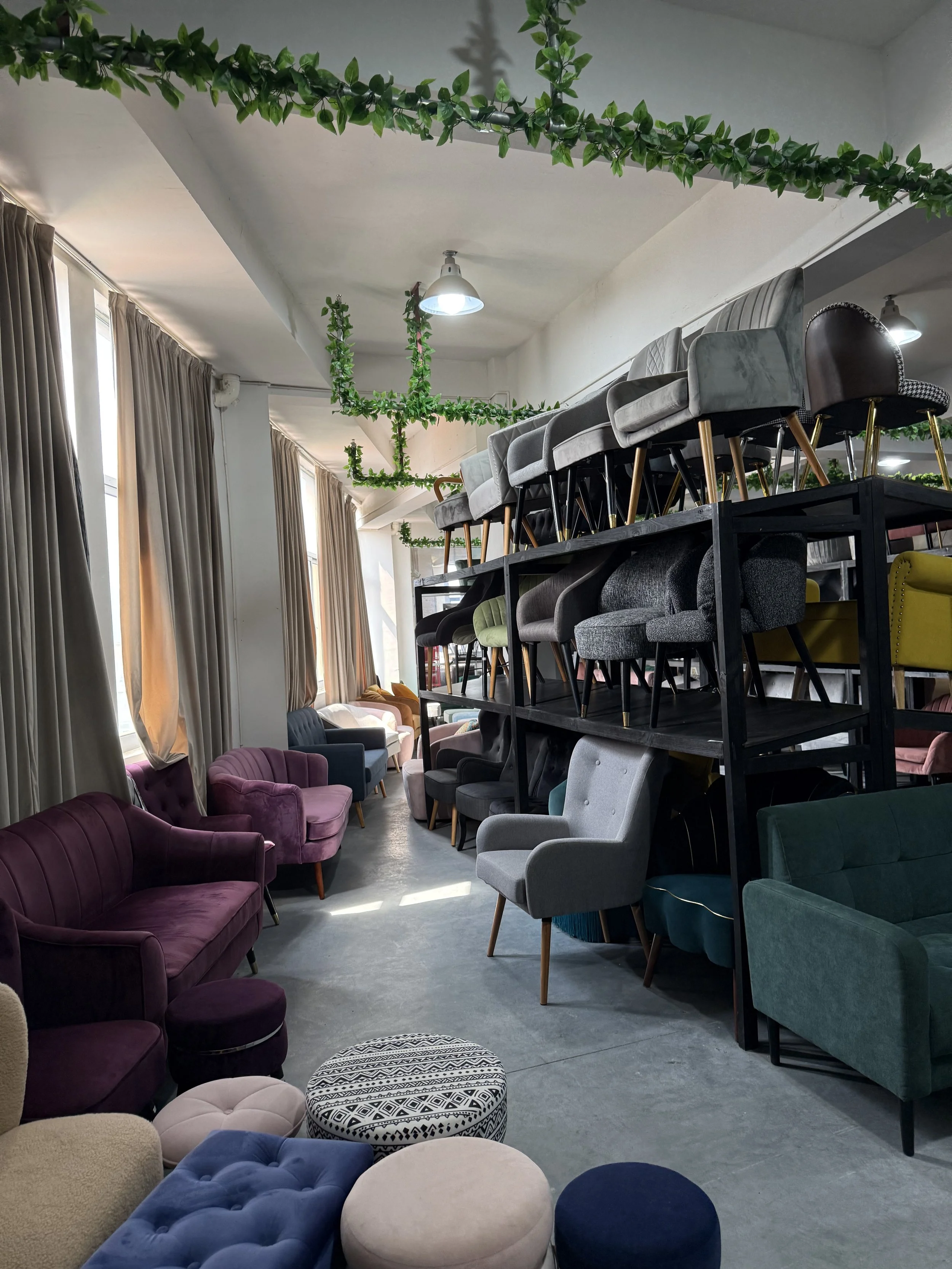 Interior of a furniture store displaying various upholstered chairs and sofas in different colors, with some stacked on shelves and others arranged on the floor. Green decorative plants hang from the ceiling and cascade down the walls. Large windows