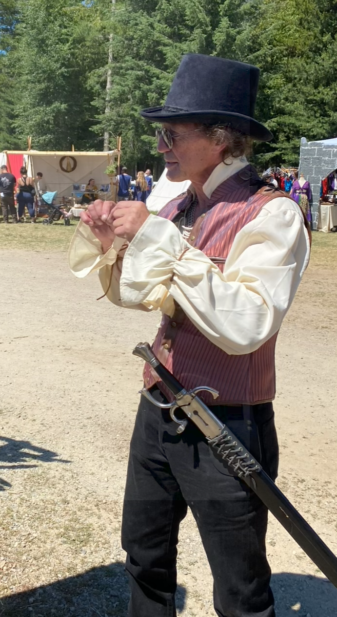 A man dressed in a steampunk costume holding a prop sword at an outdoor event with tents and people in the background.