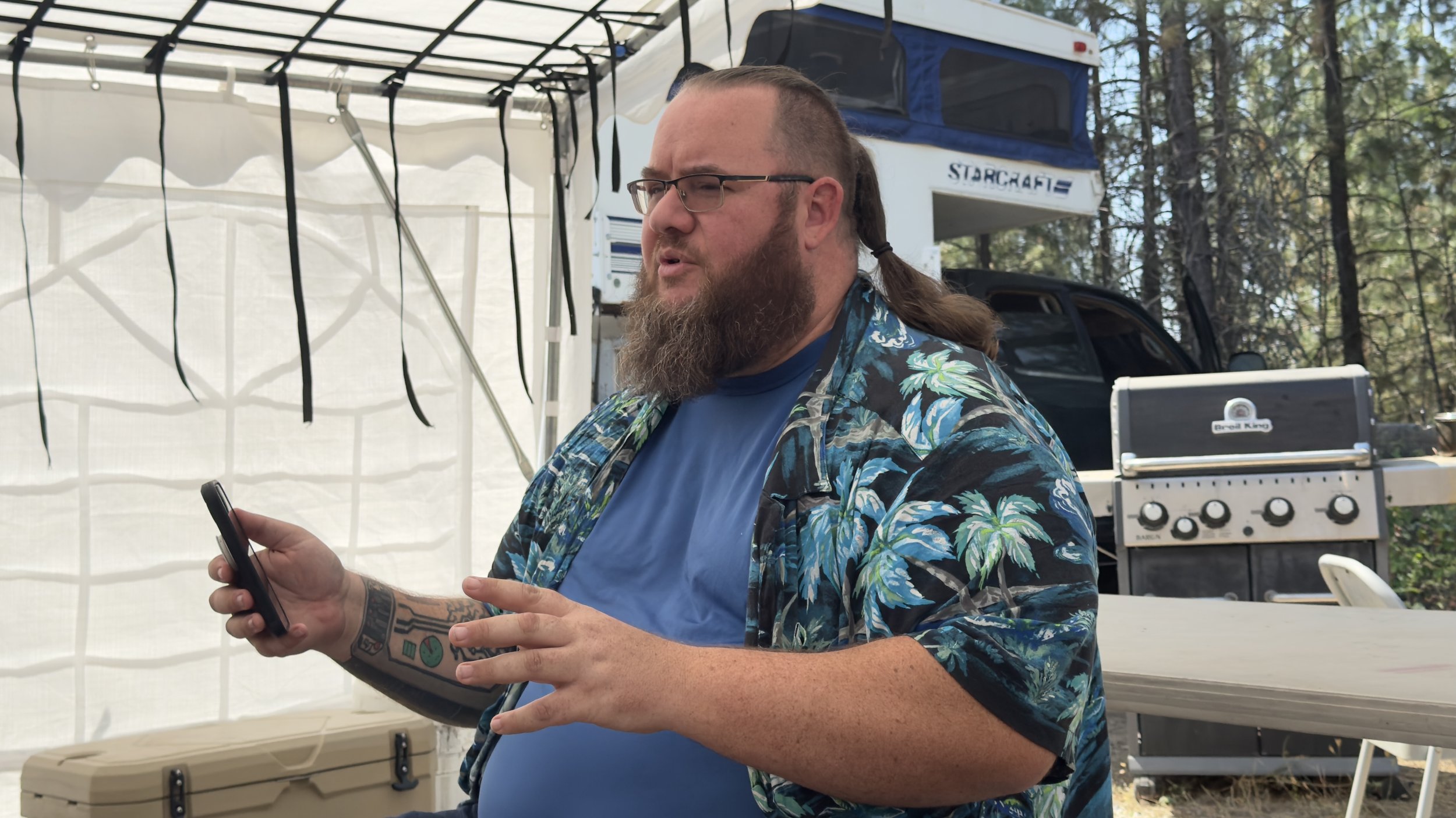 A man with a beard, glasses, and long hair tied in a braid during daytime. He is wearing a blue t-shirt and a tropical patterned shirt, holding a smartphone in his right hand and gesturing with his left. Behind him, there is a white canopy, a black t