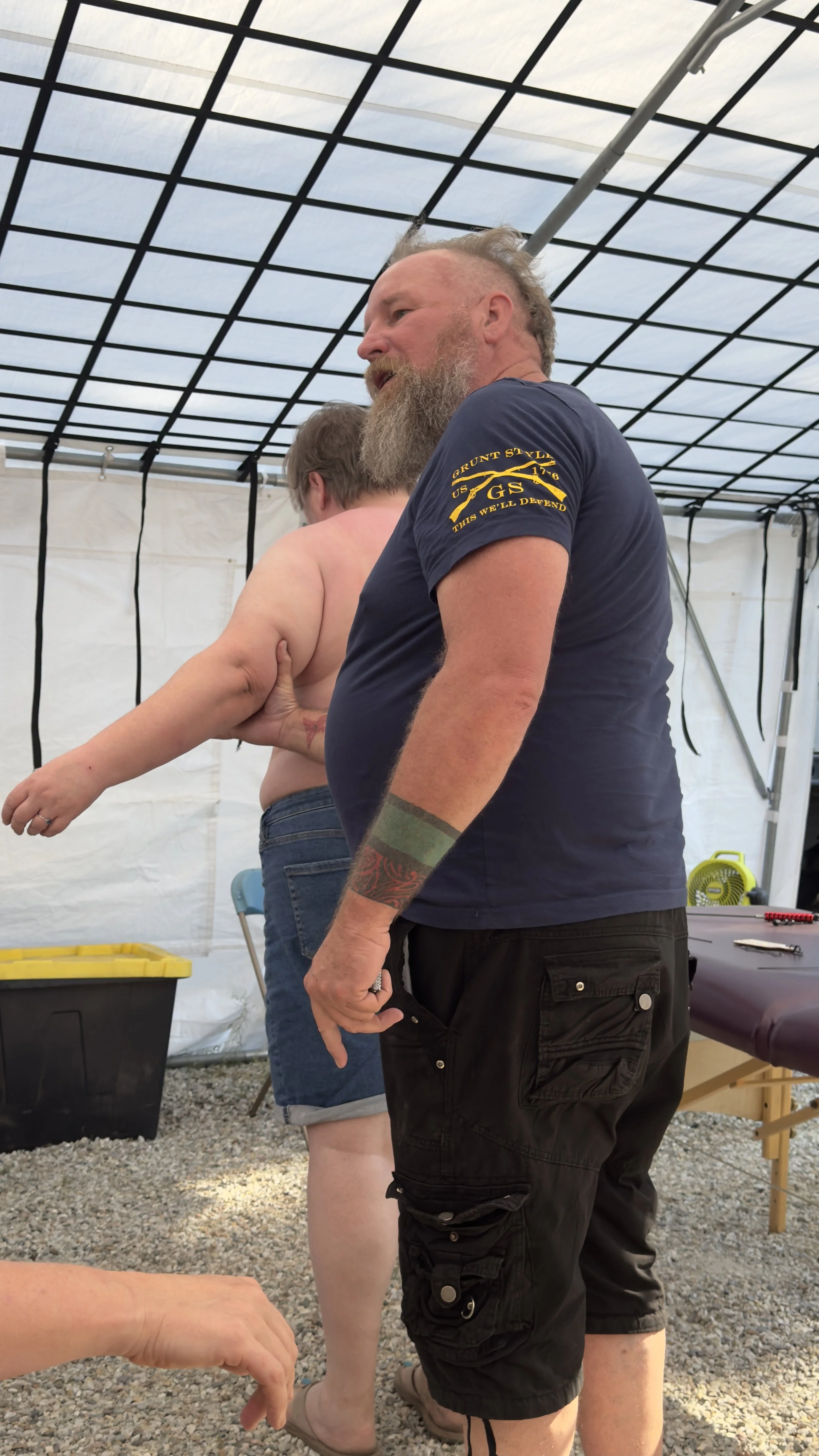 A shirtless man with glasses and a tattoo on his left arm, standing with his back to the camera. An older man with a beard, wearing a dark blue shirt with yellow text on the sleeve, stands next to him inside a tent. The scene appears to be a training