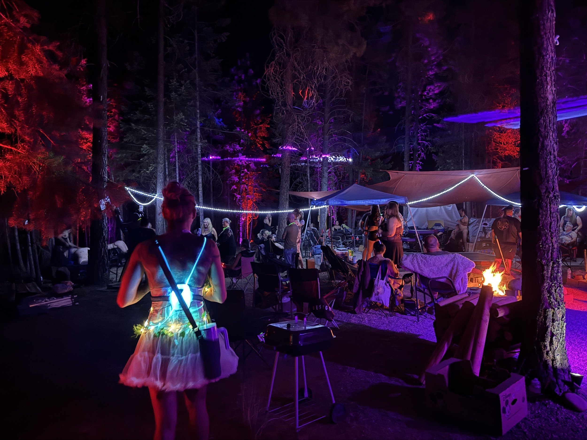 Nighttime outdoor gathering in a forest with colorful string lights, tents, people sitting around a fire, and a woman in a tutu dress with glow accessories.
