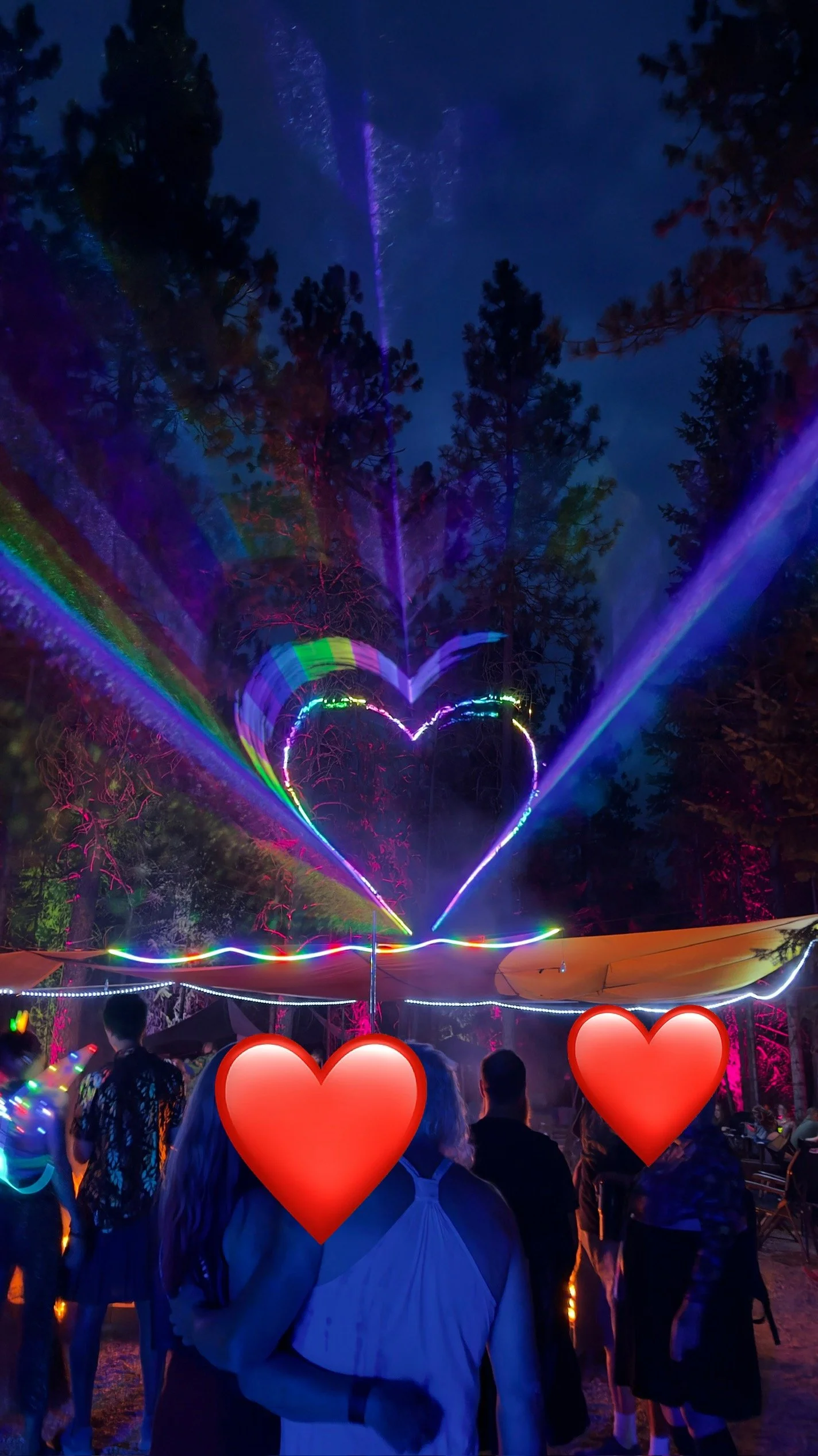 Nighttime outdoor party with colorful laser lights forming a heart shape in the sky, surrounded by trees and people dancing.