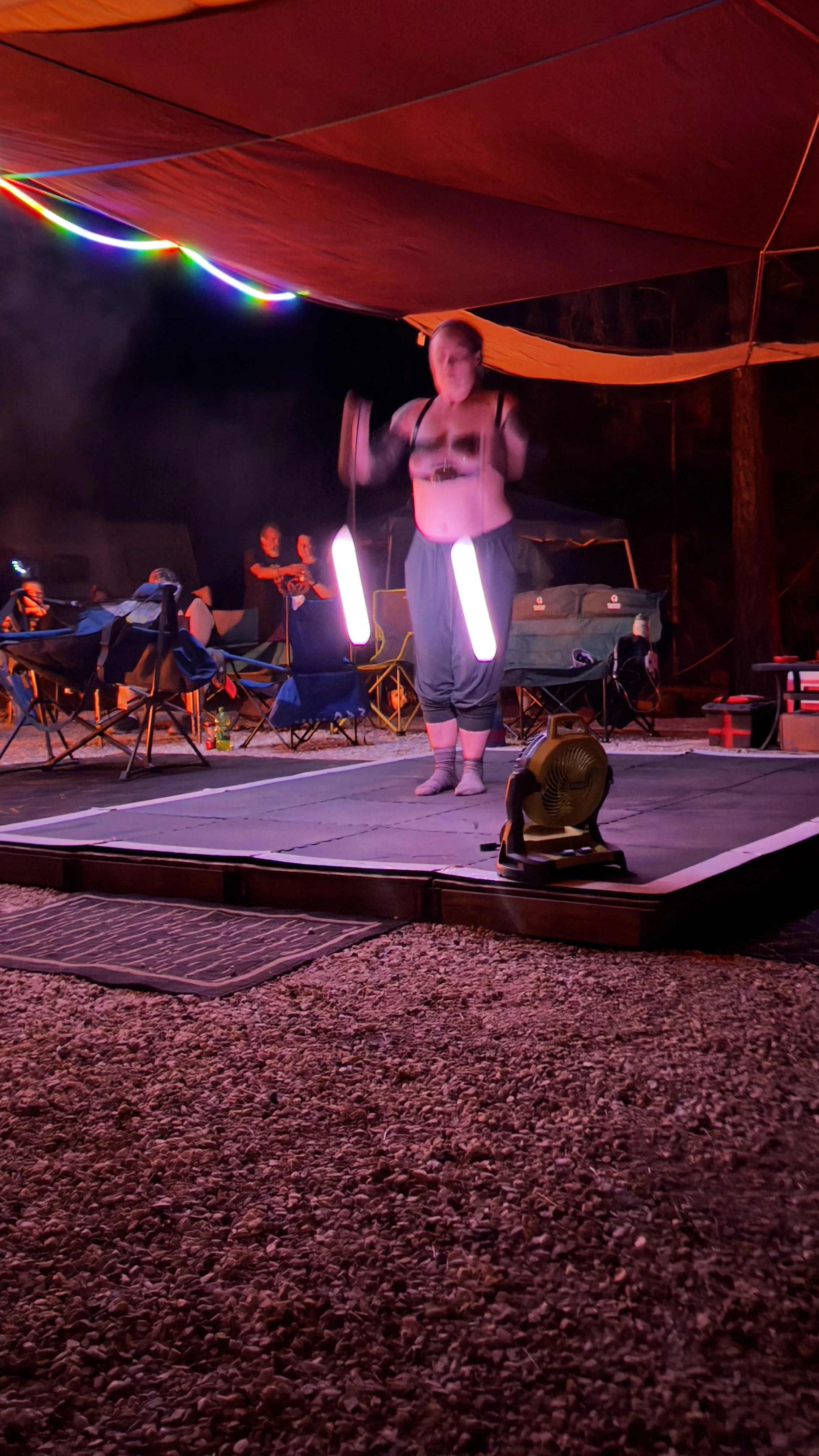 A performer on a small stage at night, illuminated by colorful LED lights, with an audience seated nearby.