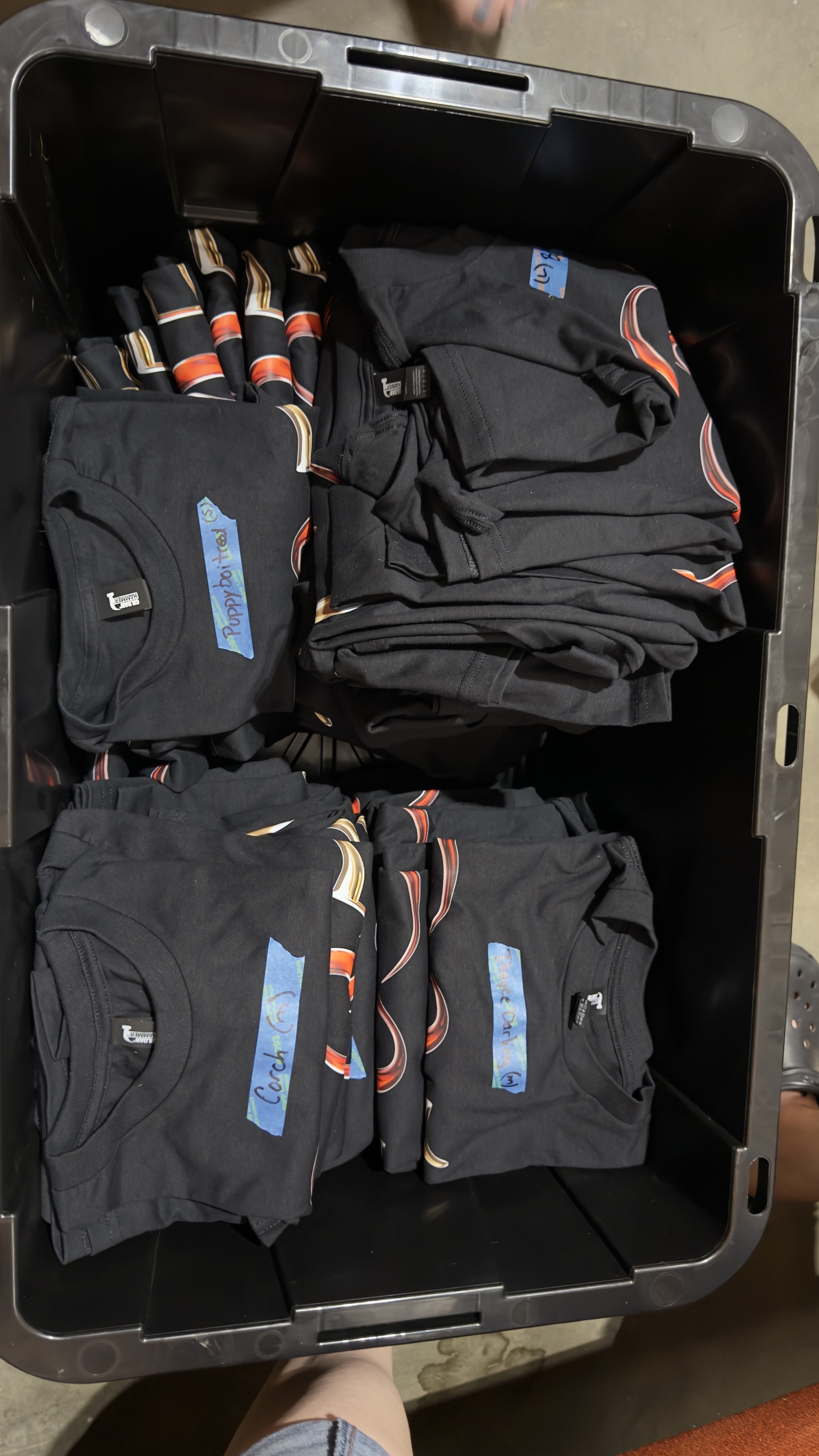 Black T-shirts with colorful designs and blue handwritten labels for different coaching groups inside a black plastic storage bin.