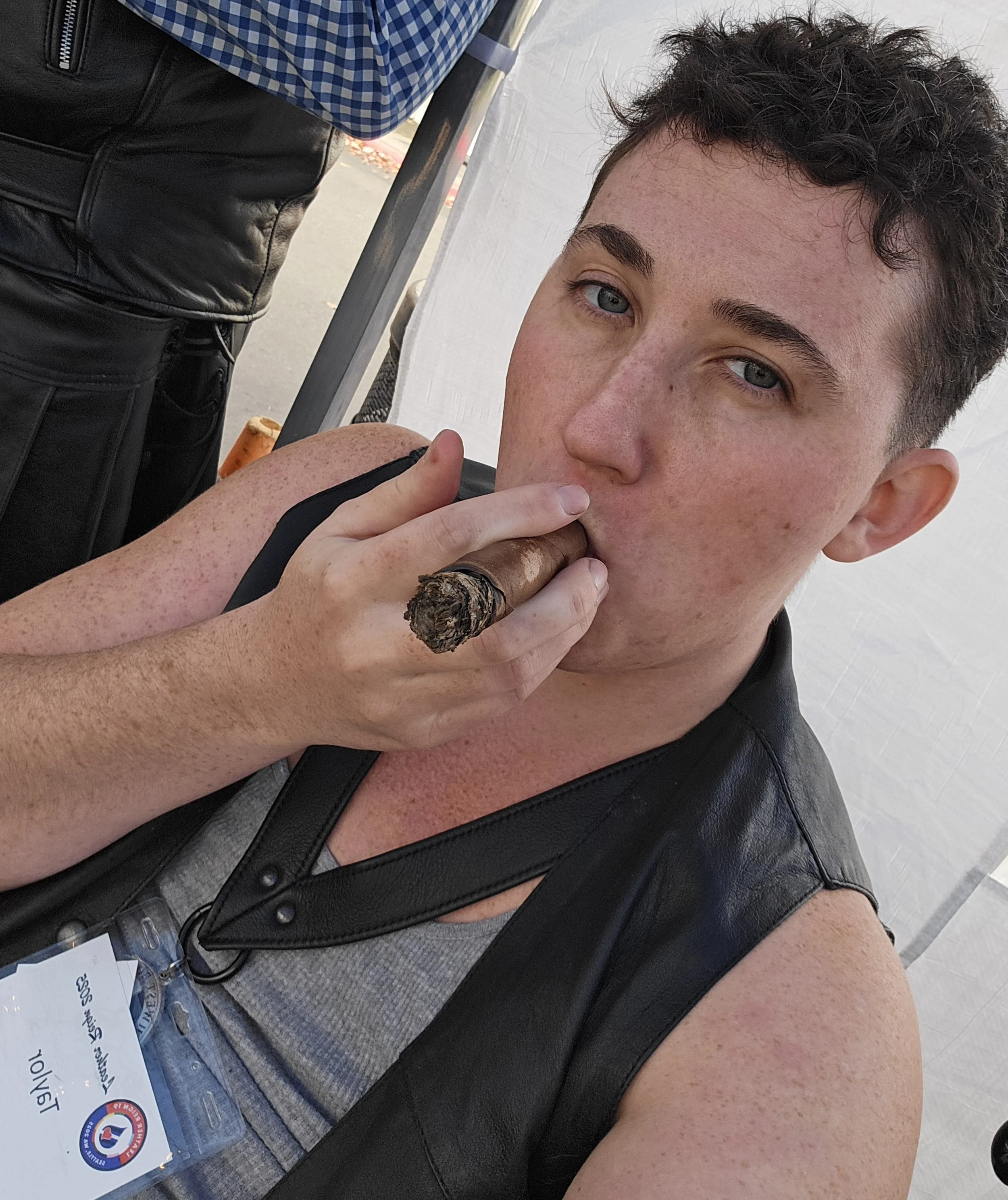 A young man with short dark curly hair and blue eyes smoking a cigar. He is wearing a gray tank top and a black leather vest. There is a name badge hanging from his neck. The background shows part of a person wearing black leather pants and a blue checkered shirt, and an outdoor setting.