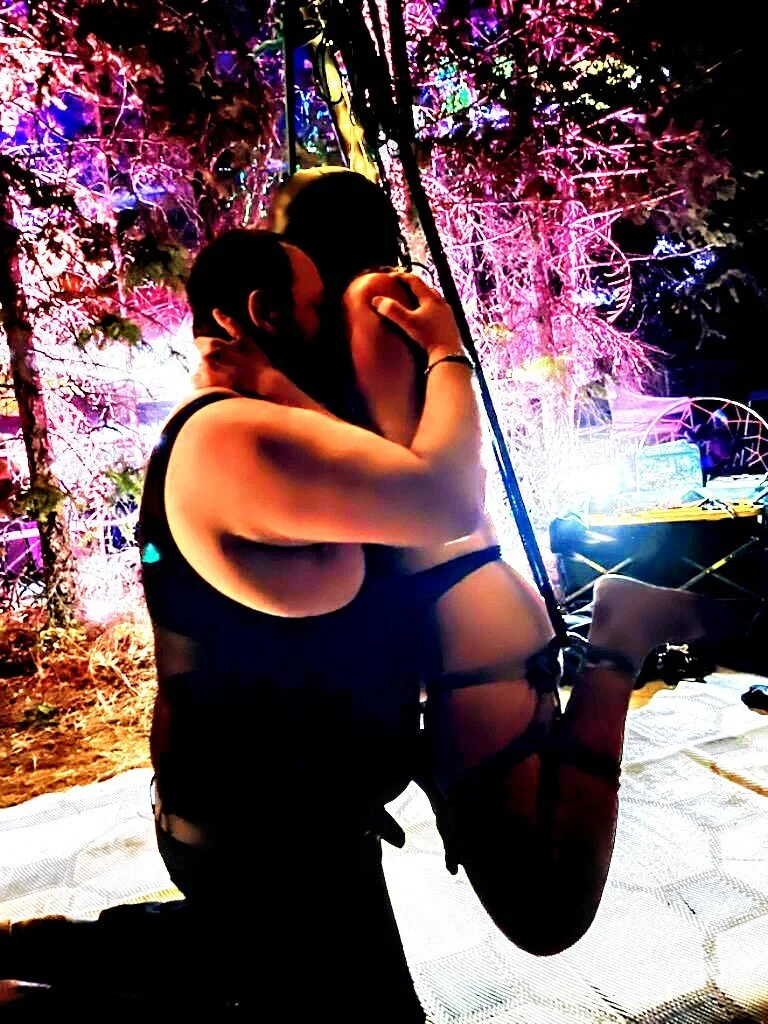 A couple is embracing and kissing in a colorful, outdoor setting with purple, pink, and blue lighting, surrounded by trees and illuminated foliage.