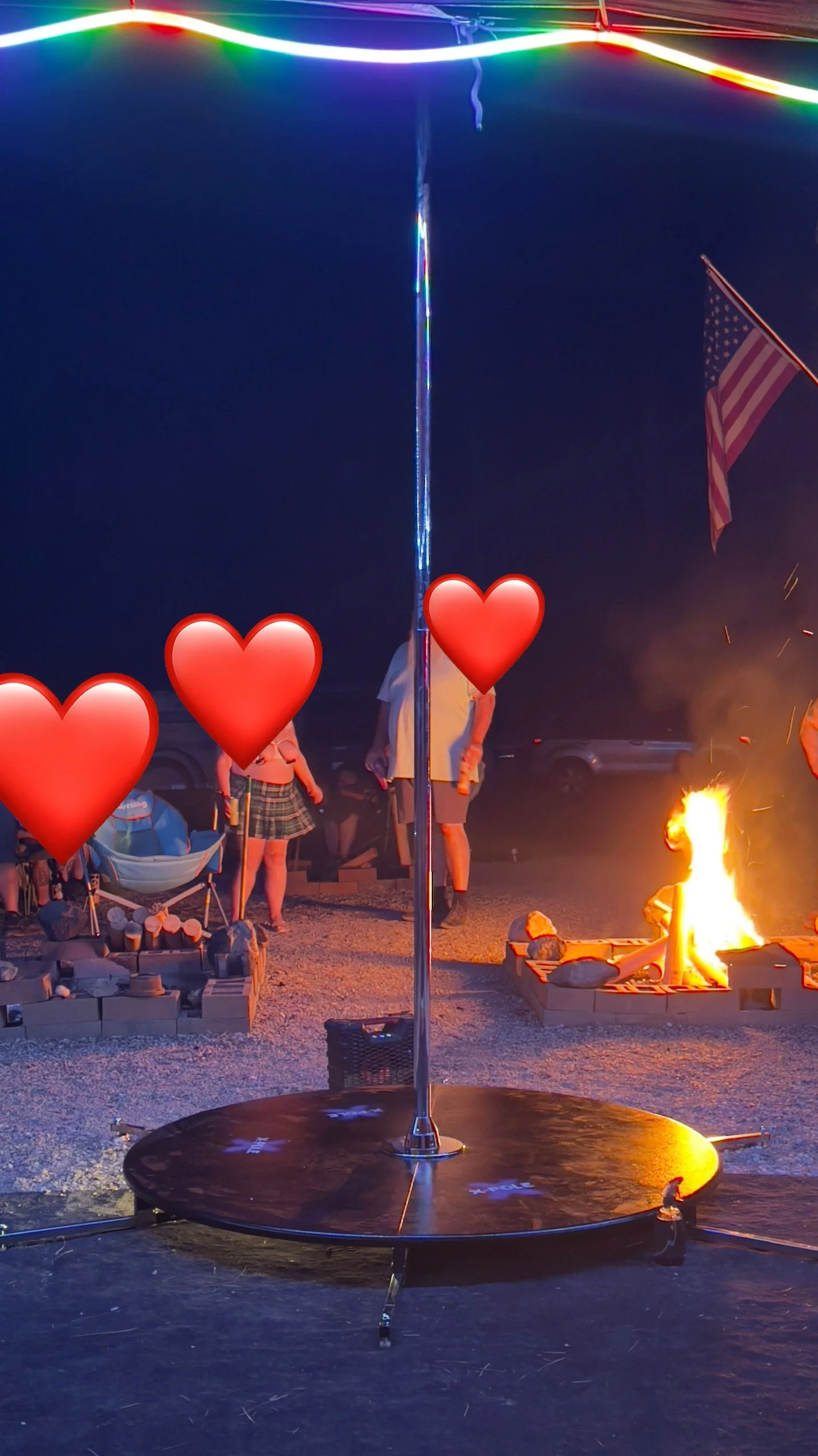 Nighttime outdoor scene with a small round platform, a vertical pole in the center, and a fire pit with a person sitting near it. Several people are gathered in the background, and an American flag is visible. Colorful neon lights are overhead, and t