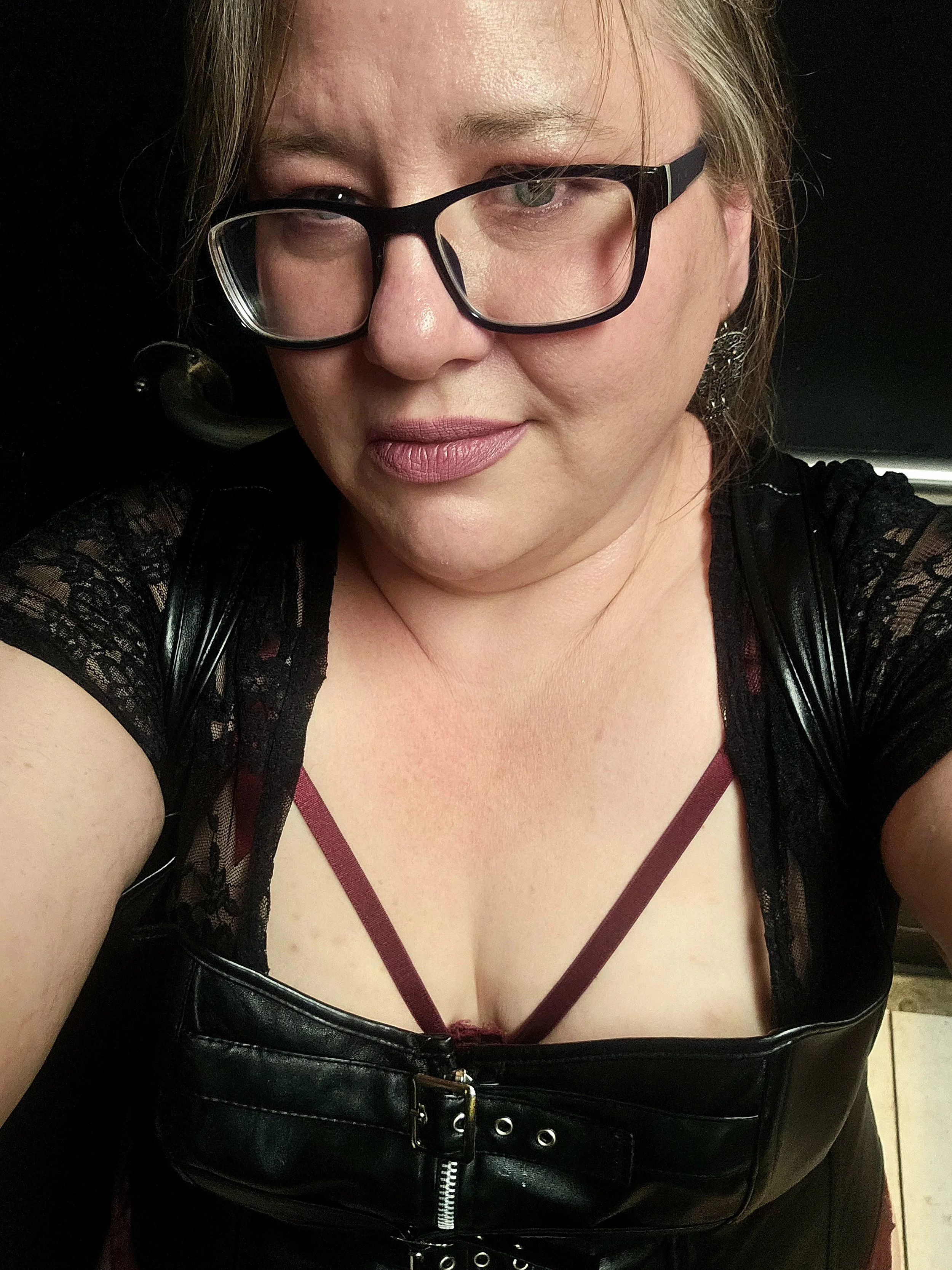 A woman with glasses, wearing a black lace top and a black leather outfit, taking a selfie.