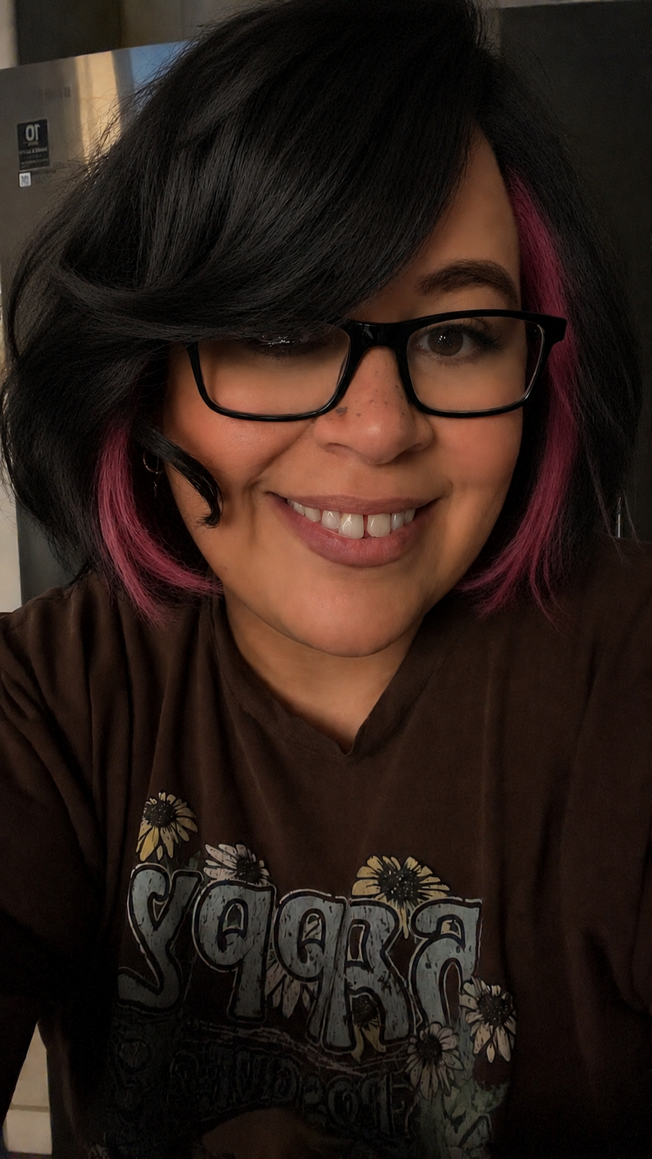 A woman with black hair with pink streaks, glasses, and a brown t-shirt with floral and text print, smiling at the camera.