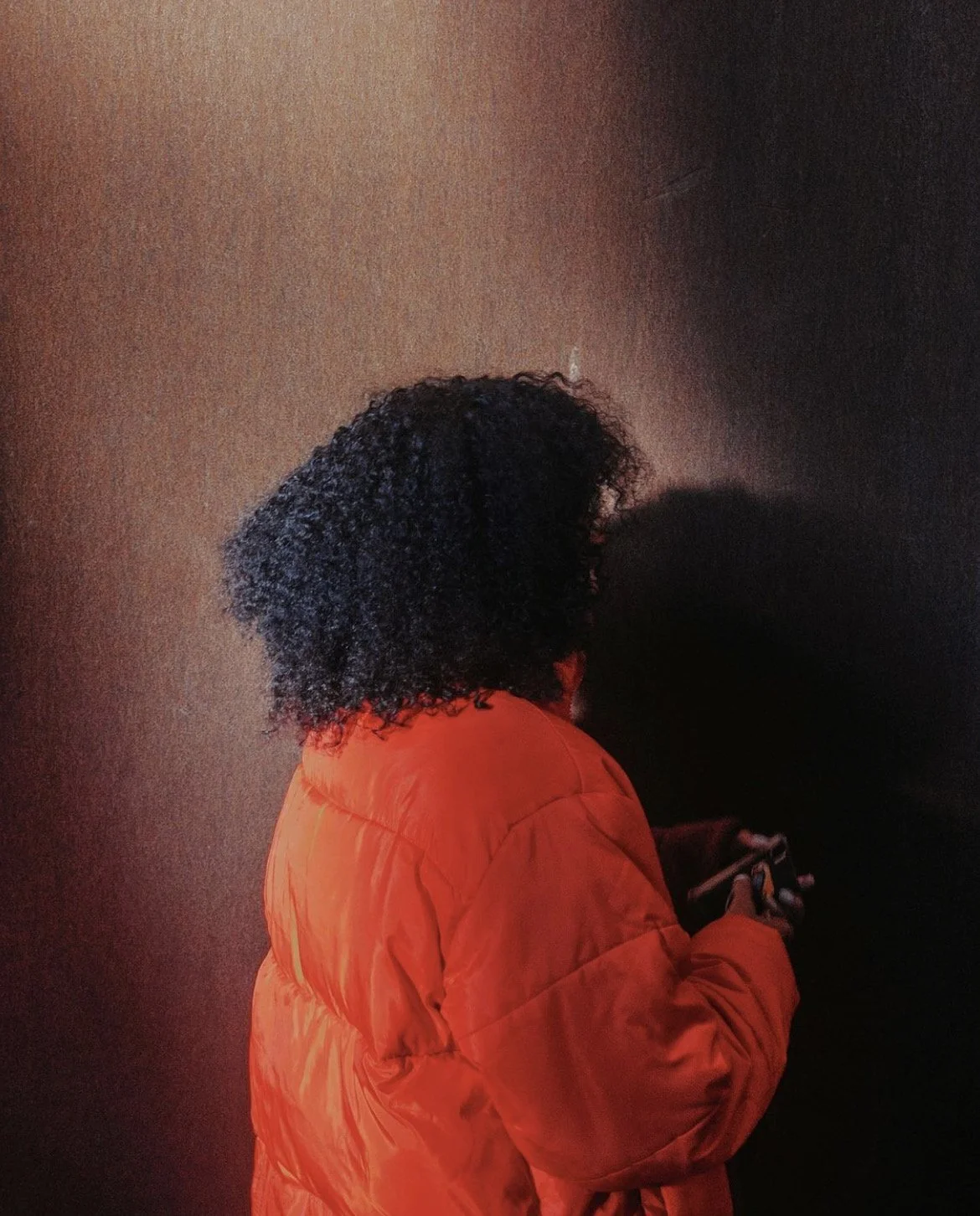 Person in orange jacket with curly hair using a cellphone against a dark background.