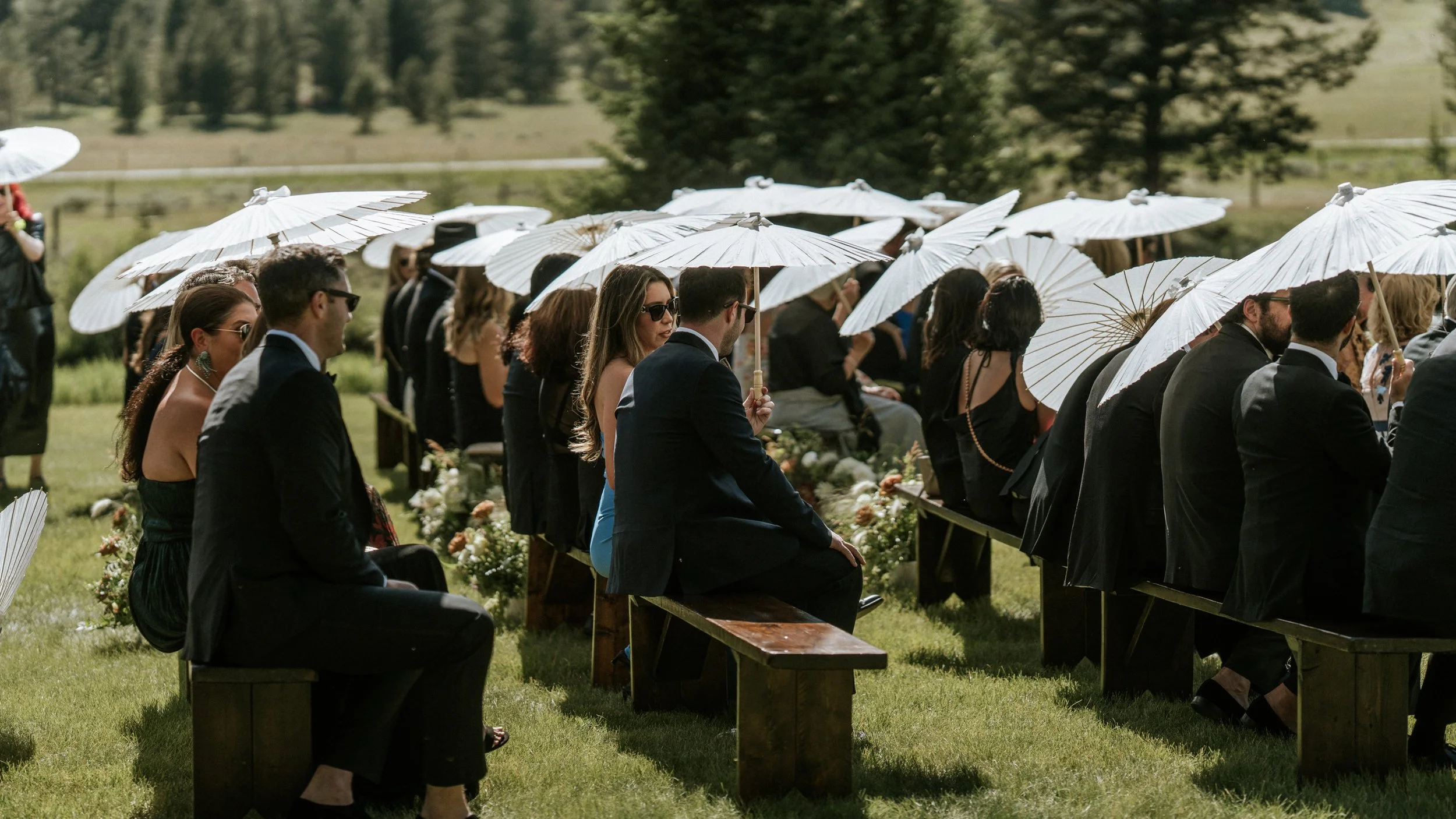 People attending an outdoor wedding ceremony, sitting on benches in a grassy field, holding white parasols for shade, dressed in formal attire, with trees in the background.