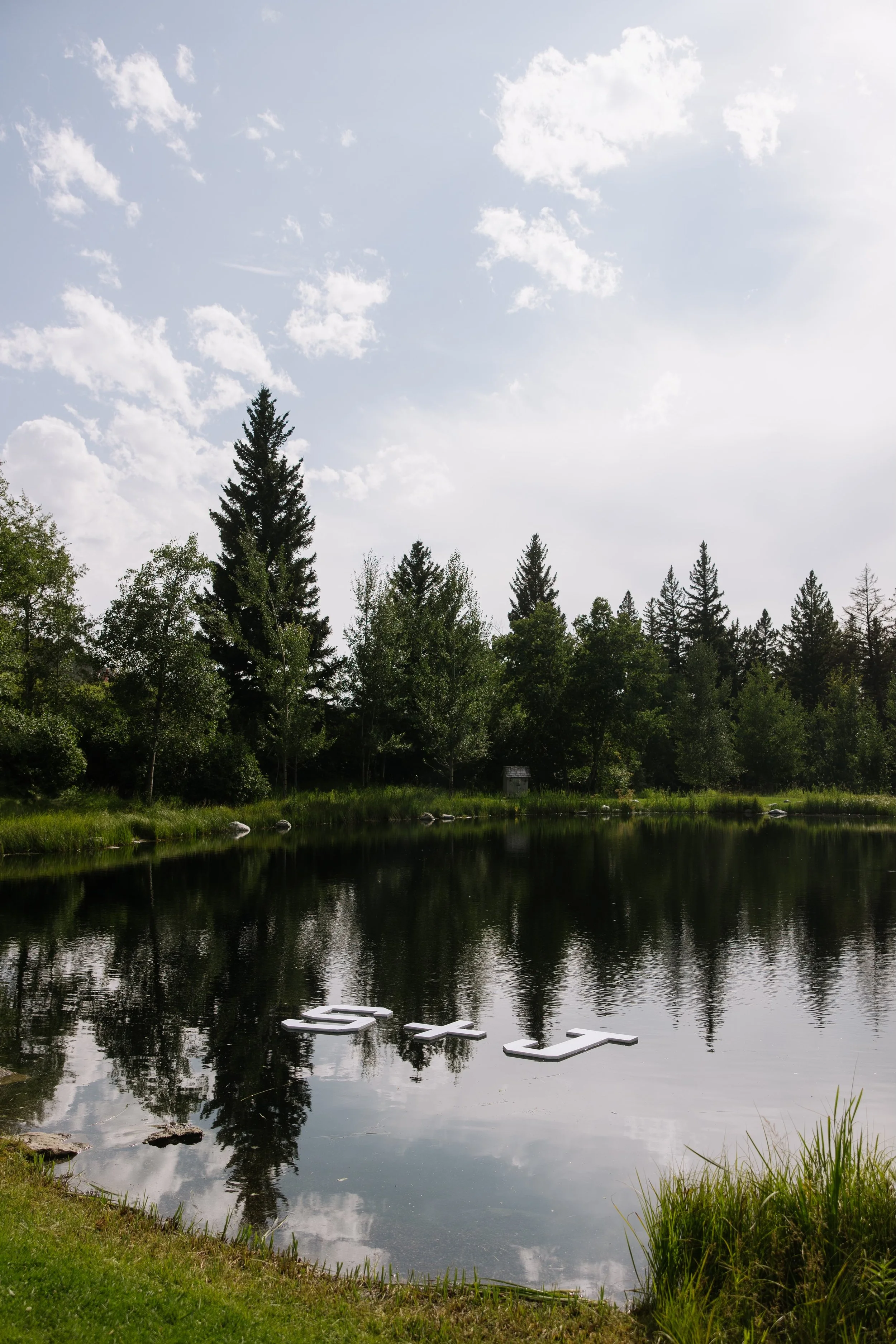 Serene lake with reflective water surrounded by green trees and grass, with white floating letters spelling 'HELLO' on the surface and a partly cloudy sky above.