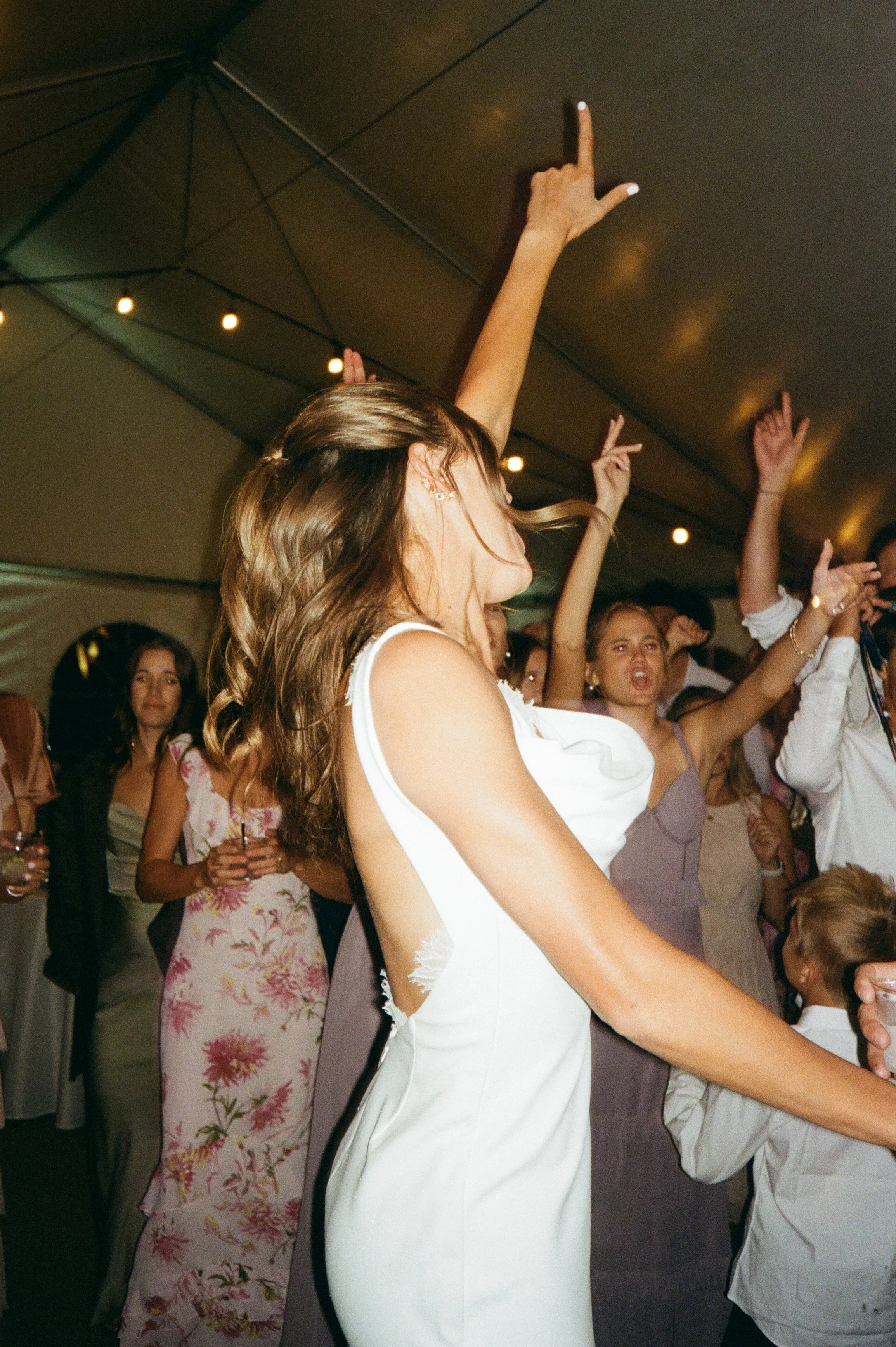 People dancing and celebrating at a party, women in dresses and children in a large indoor tent with string lights.