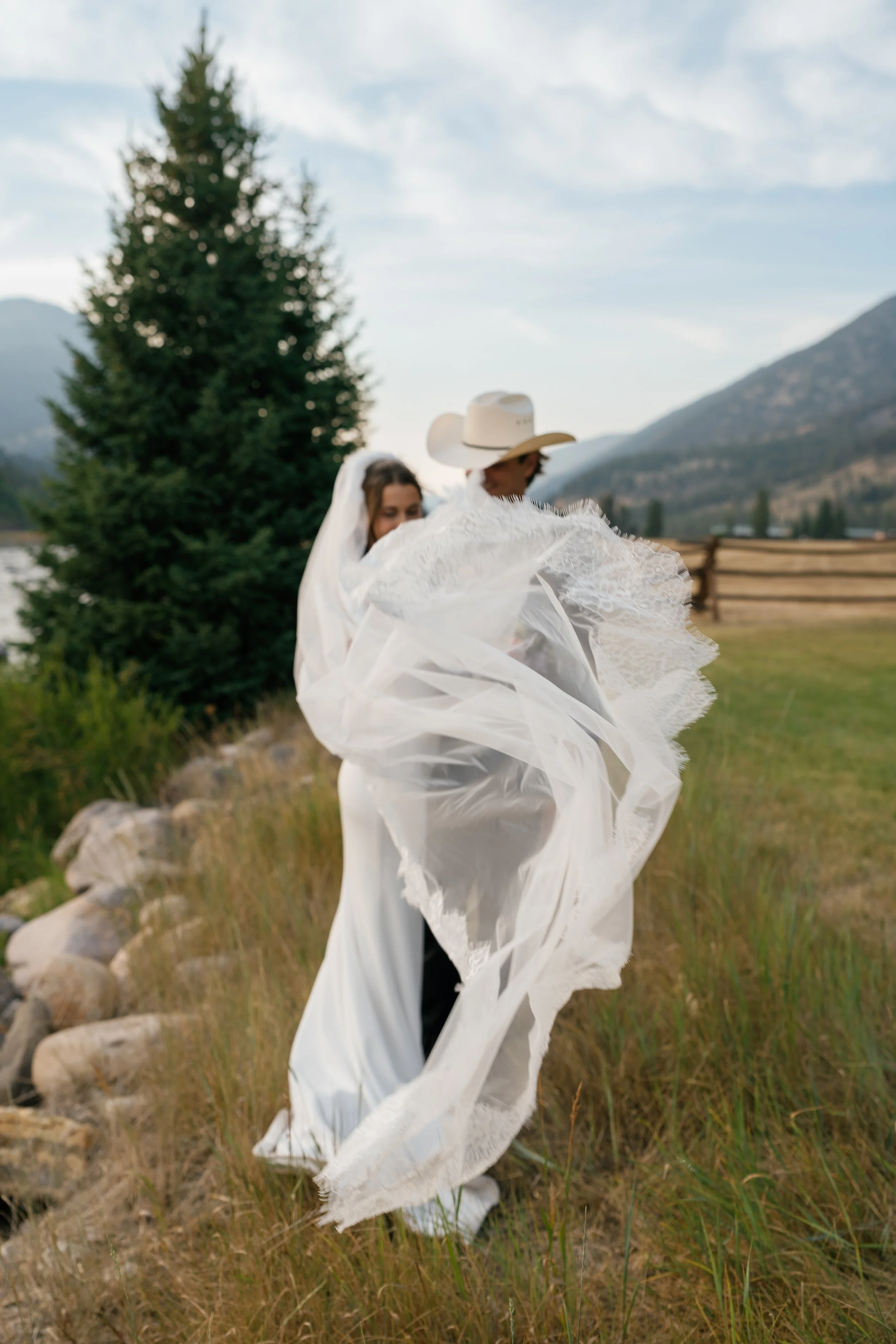A bride and groom outdoors, with the groom wearing a cowboy hat, standing on grassy terrain near a stone-lined path, with mountains and trees in the background, as the bride’s veil flows in the wind.