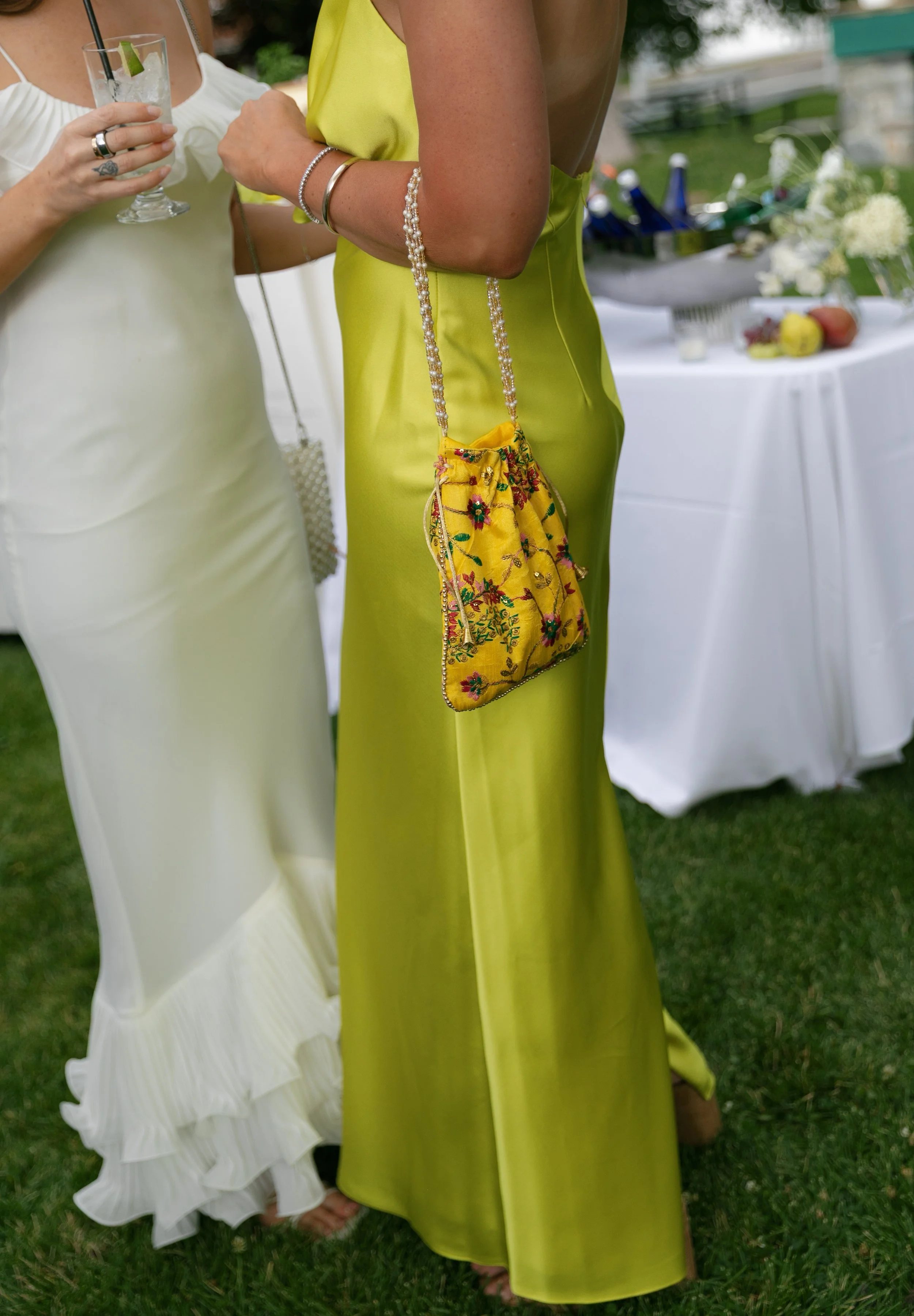 Two women in formal dresses, one in a white dress with ruffled details at the bottom, the other in a bright yellow satin dress, holding a yellow embroidered handbag with a beaded strap, at an outdoor event with a table of food and flowers in the background.