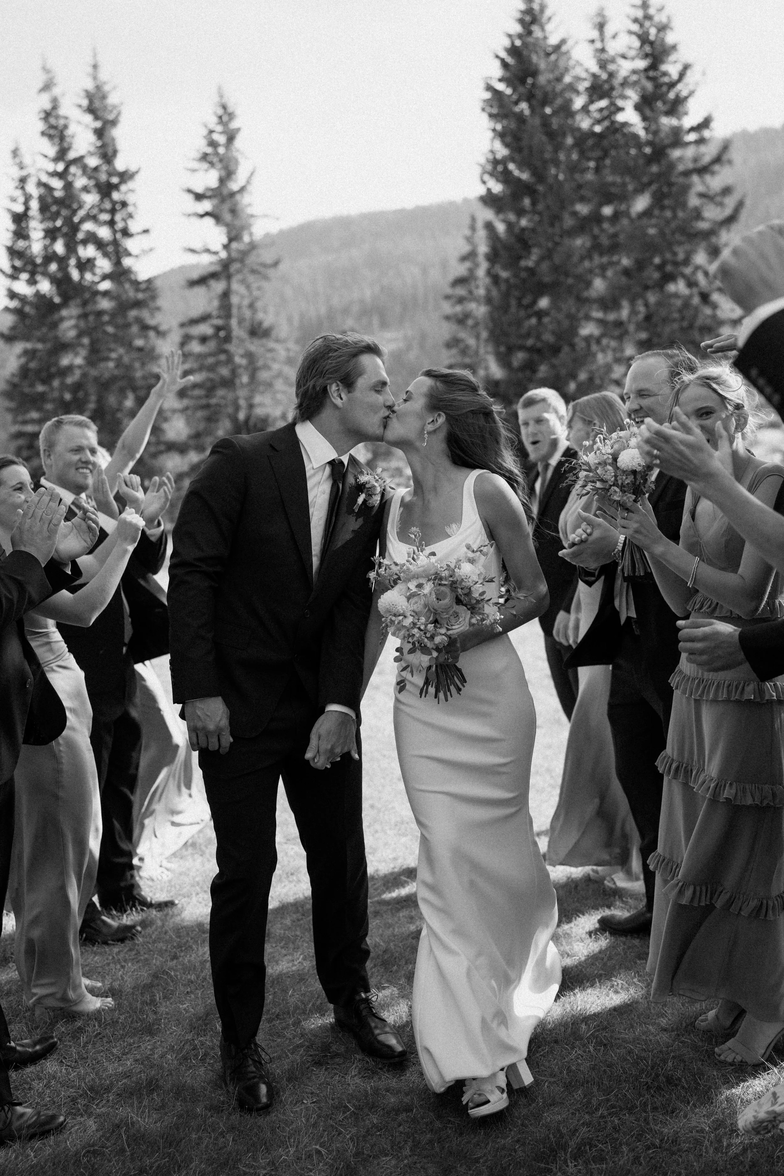 A black-and-white photo of a wedding scene where a couple is kissing, surrounded by friends and family clapping and celebrating outdoors with trees and mountains in the background.