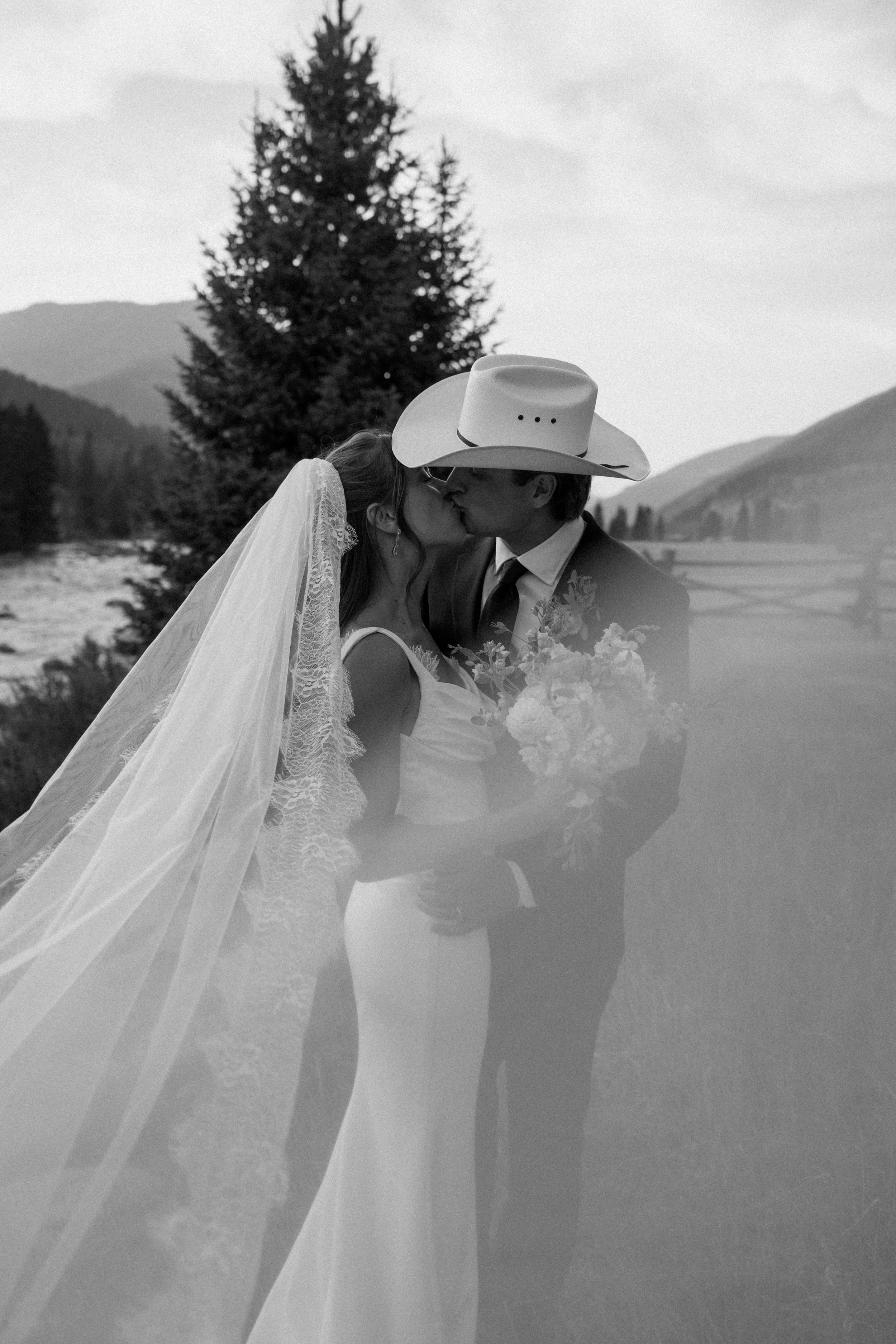 A black-and-white photo of a bride and groom sharing a kiss outdoors. The bride wears a lace wedding dress and veil, and the groom wears a suit with a cowboy hat. The bride holds a bouquet of flowers, and trees and mountains are visible in the background.