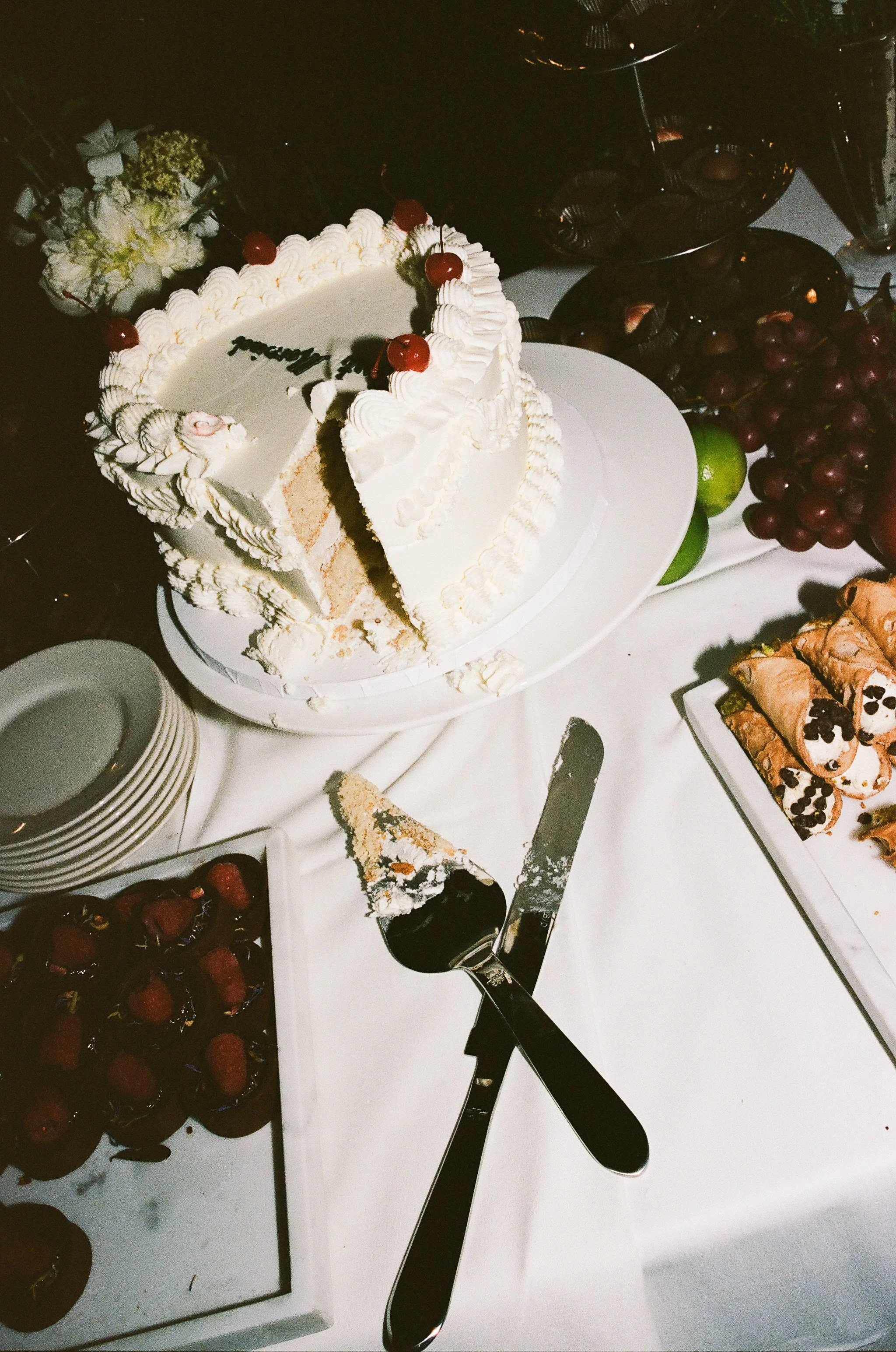 A partially sliced white birthday cake with cherries on top. Surrounding the cake are plates, a serving knife with a cake slice, strawberries in a dish, and a platter of pastries, all on a white tablecloth.