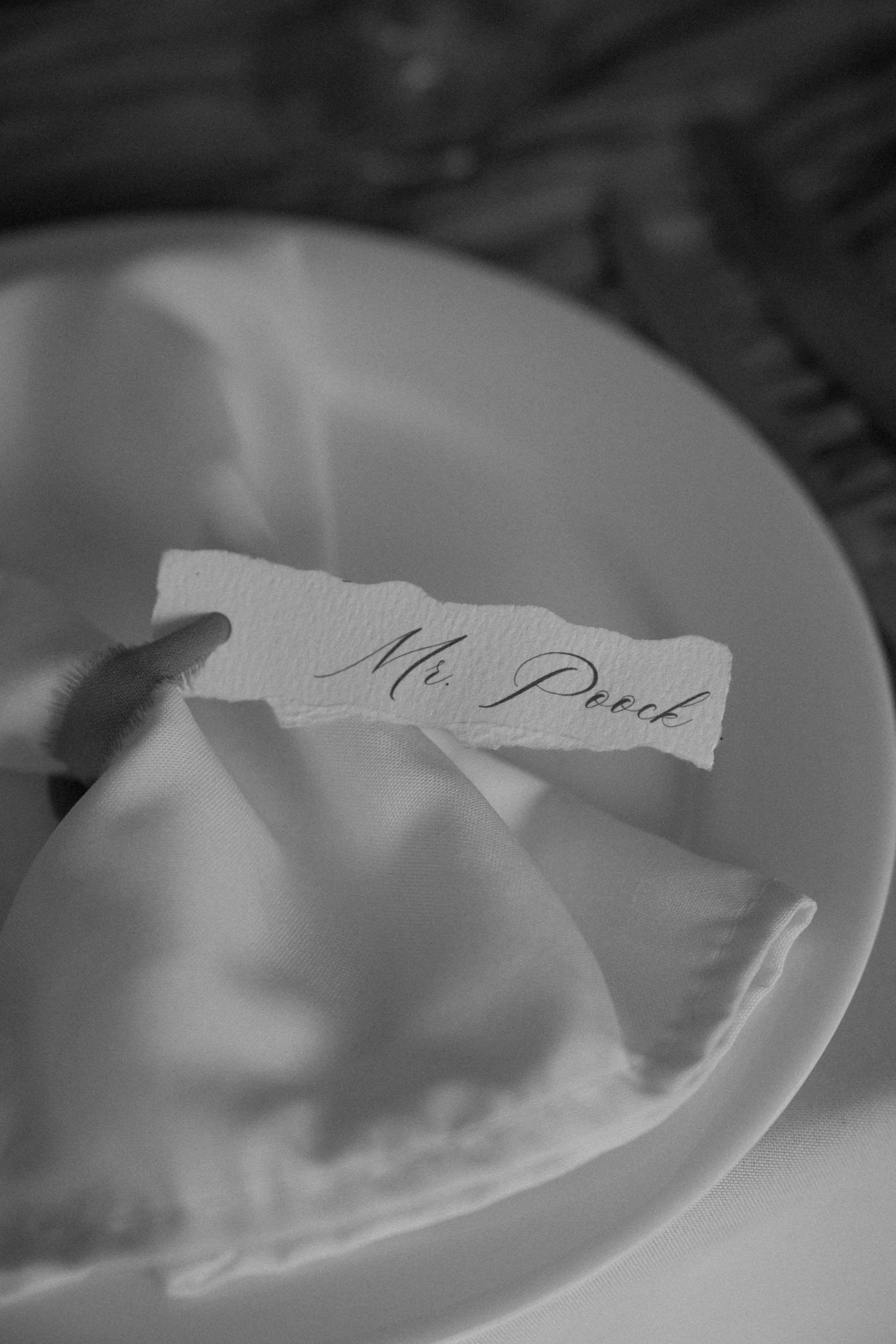 A torn piece of paper with the words 'Mr. Pock' written in elegant script, held above a satin fabric on a white plate.