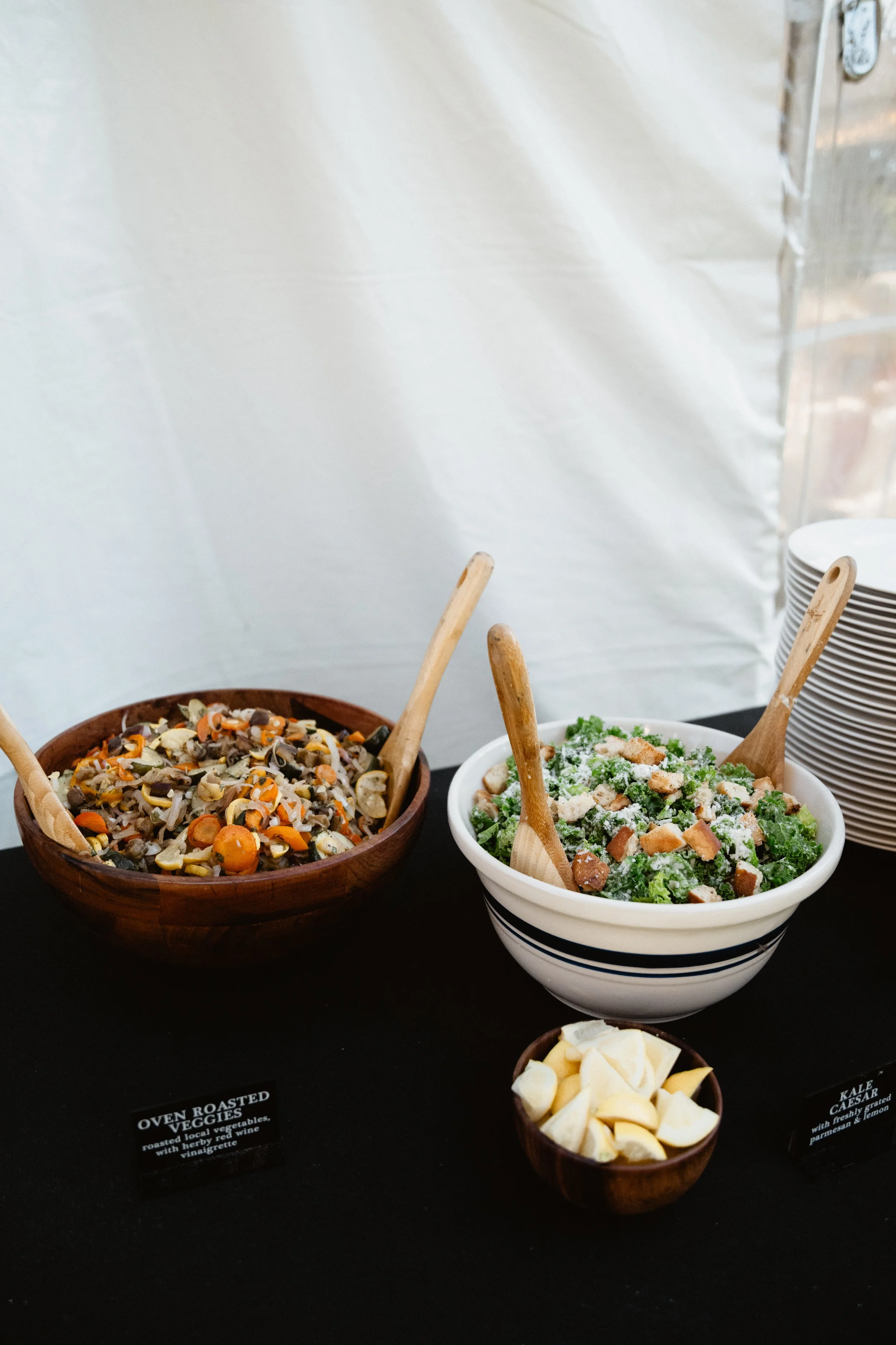 Two bowls of salad with wooden serving utensils, one containing a vegetable medley and the other a kale Caesar salad, alongside a smaller bowl of cheese, set on a black table with stacked plates nearby.