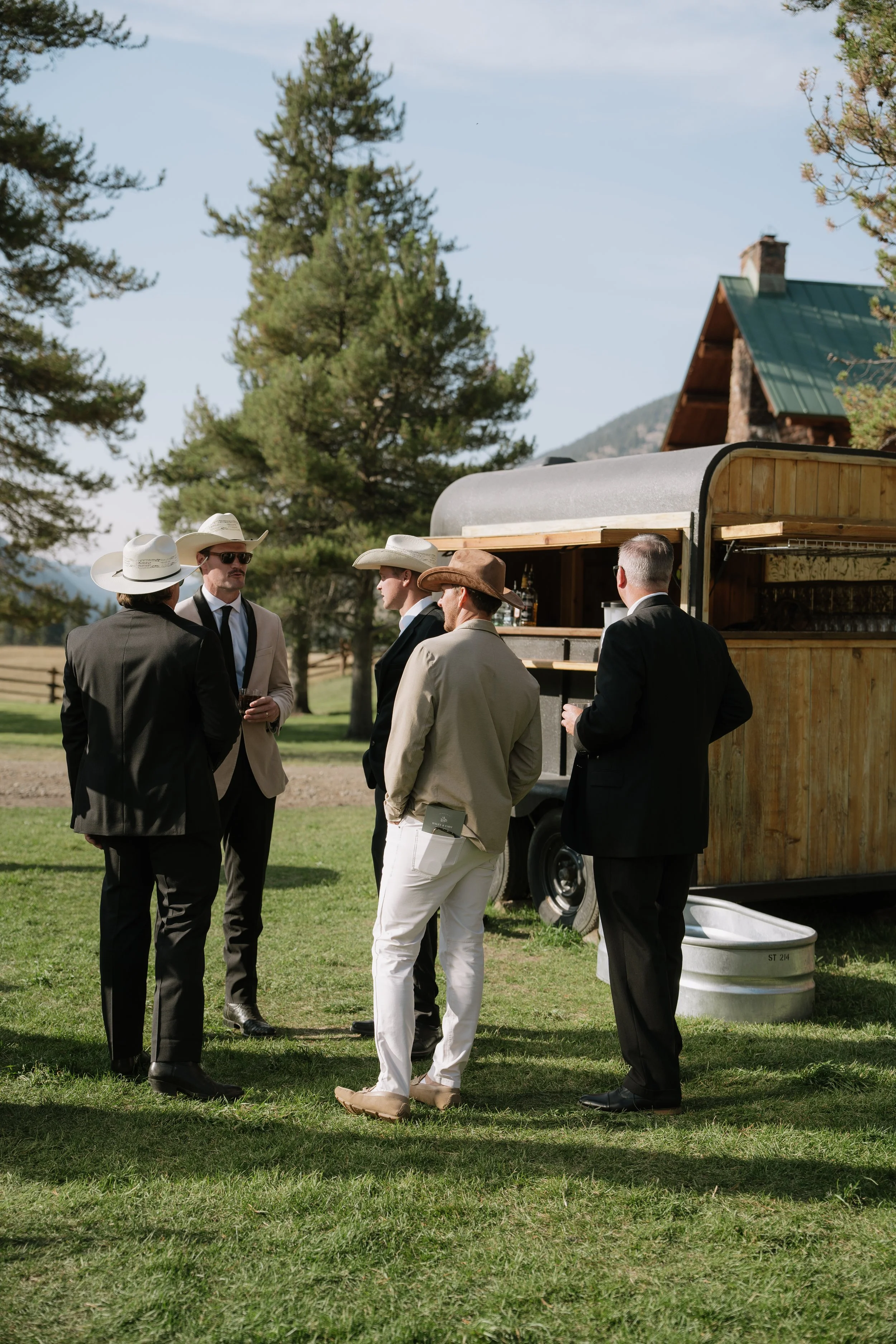 Group of men dressed in suits and cowboy hats standing outside on grass, engaged in conversation near a wooden food trailer, with trees and mountains in the background.