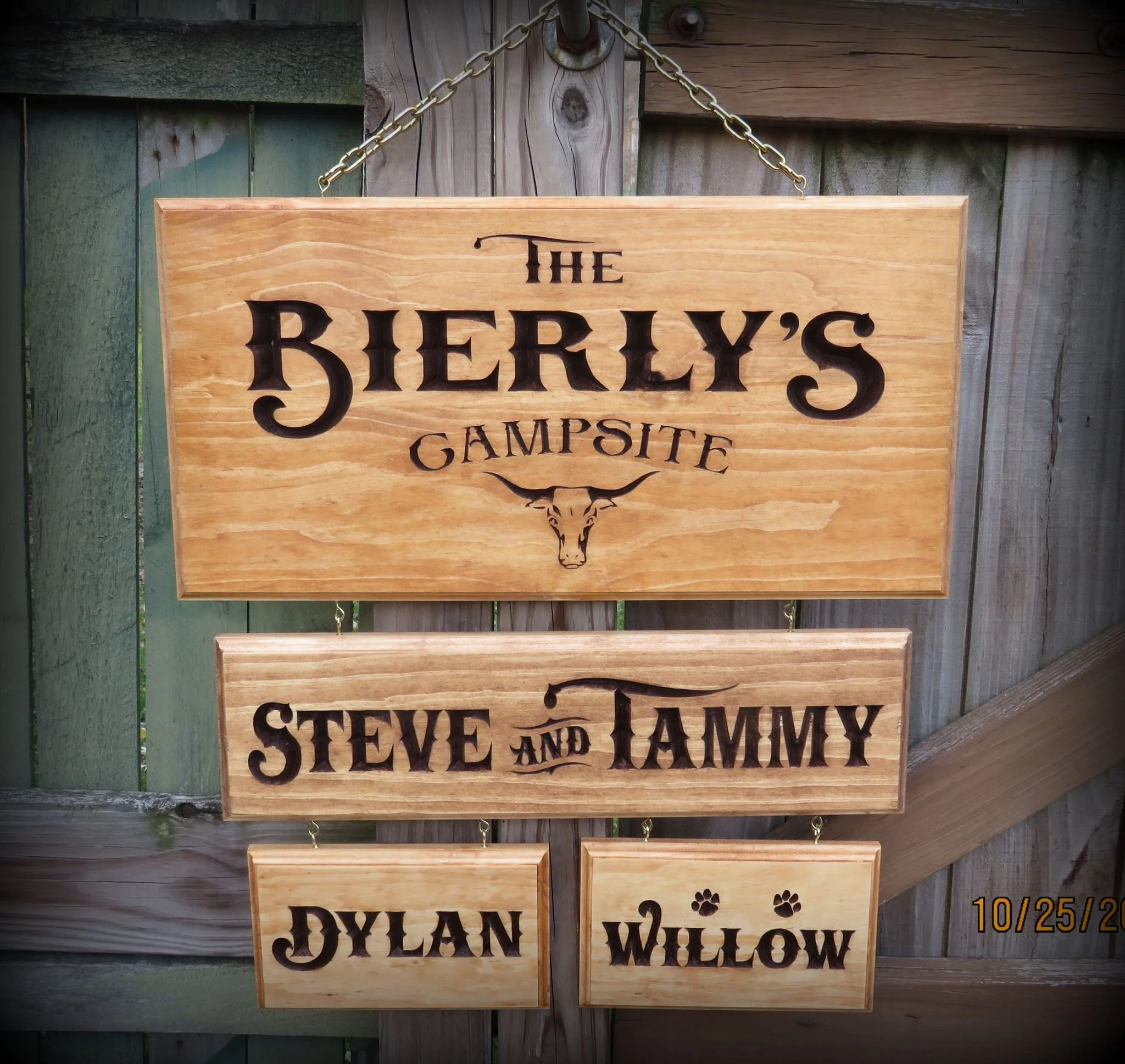 A wooden sign for 'The Bierly's Campsite' hangs on a fence, with names 'Steve and Tammy,' 'Dylan,' and 'Willow' written below, decorated with paw prints and a longhorn skull.