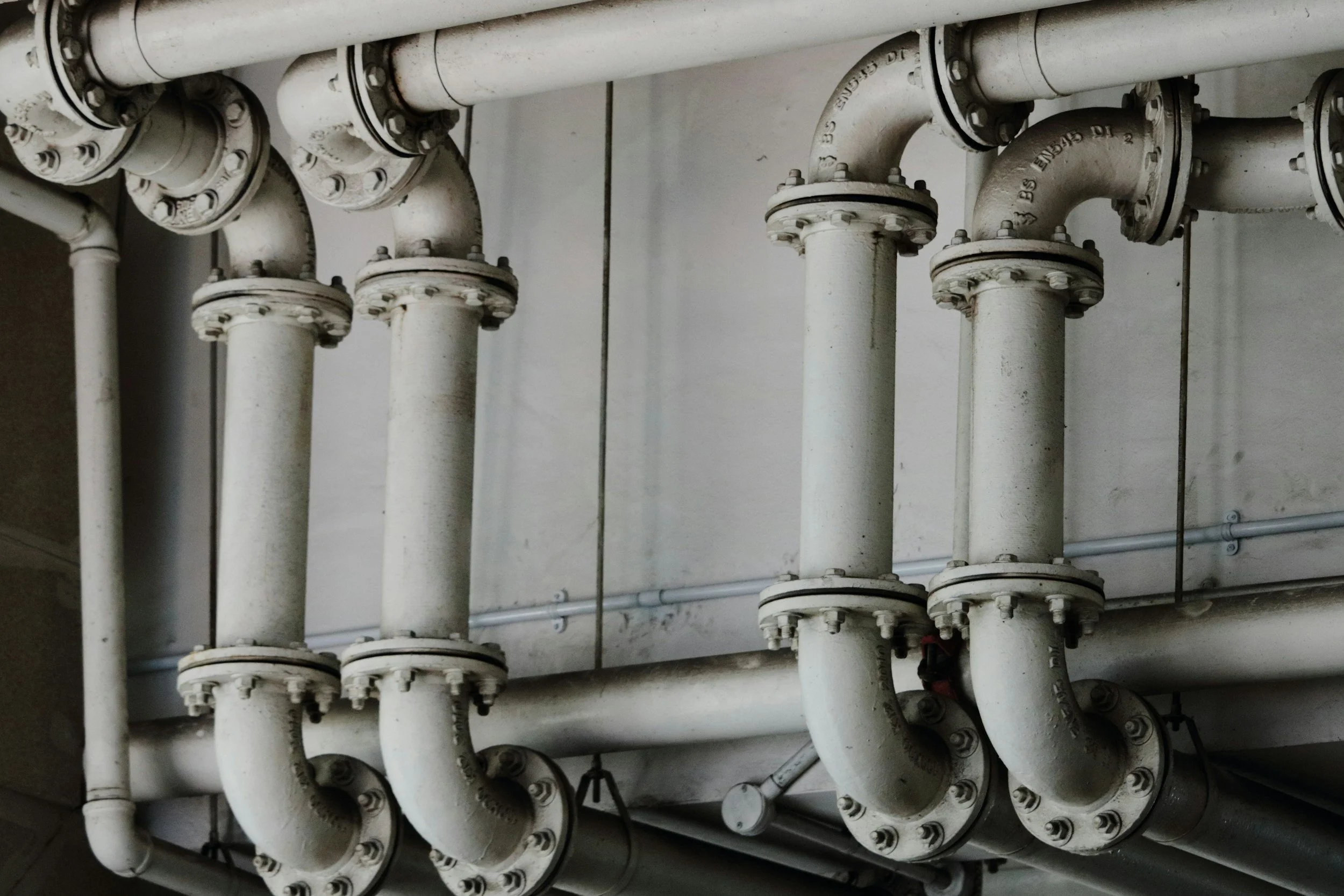 Multiple white industrial pipes with flanges and elbows running along a ceiling or wall.