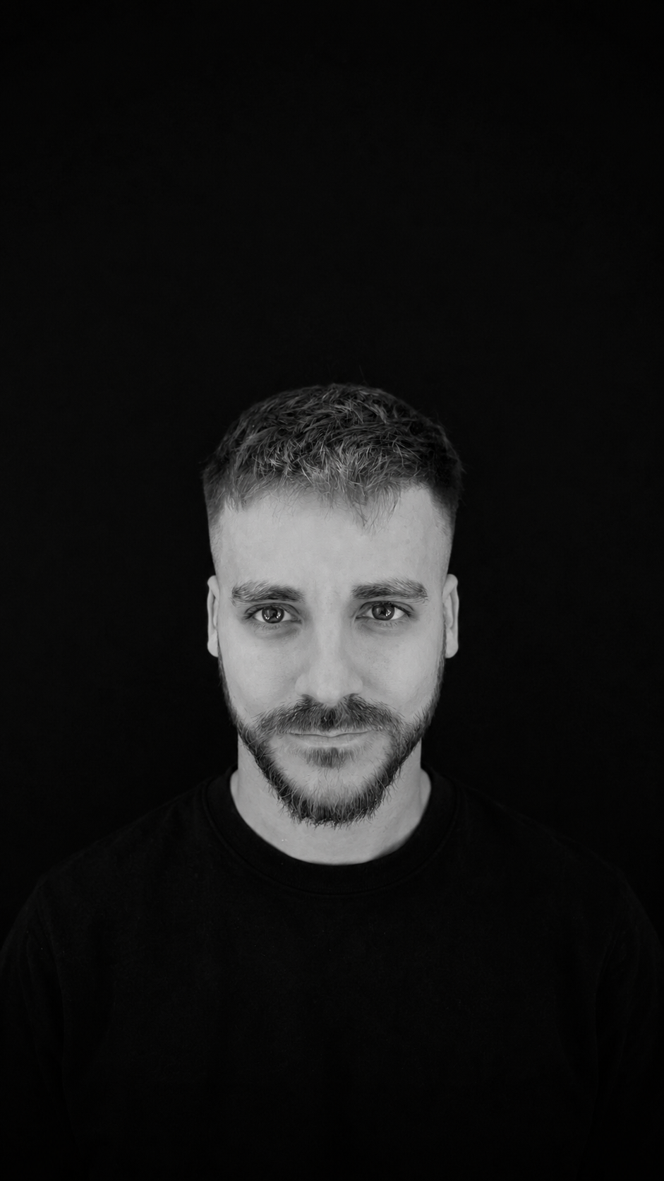 A black and white portrait of a young man with short hair and a beard, smiling slightly, against a black background.
