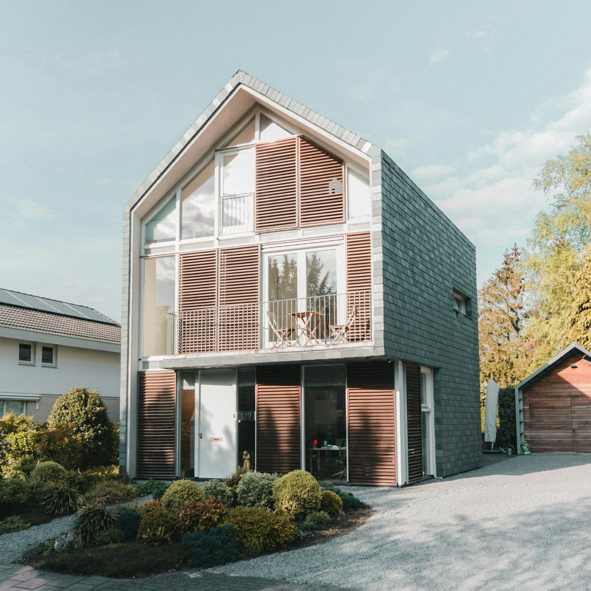 Modern three-story house with gray shingle exterior, brown wooden shutters, large glass windows and balconies, surrounded by greenery and a gravel driveway.