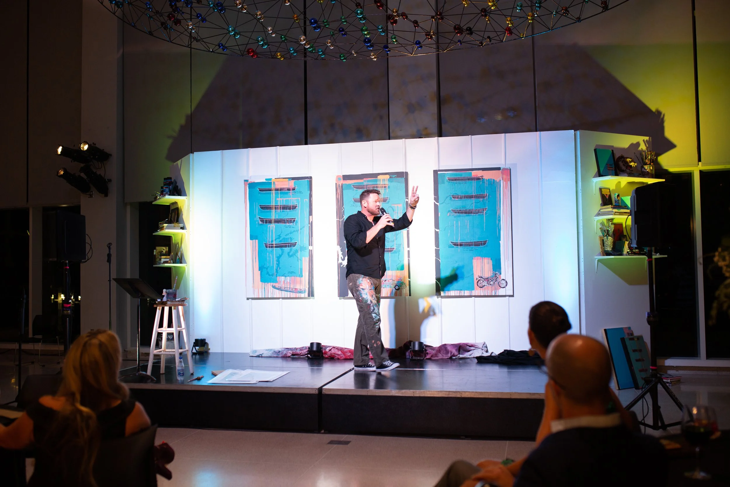 Chris McAdoo standing on the knoxville museum of art stage surrounded by his abstract paintings and colorful lights. Several audience members are seated in front of him, and a ceiling with a decorative sphere is visible above.