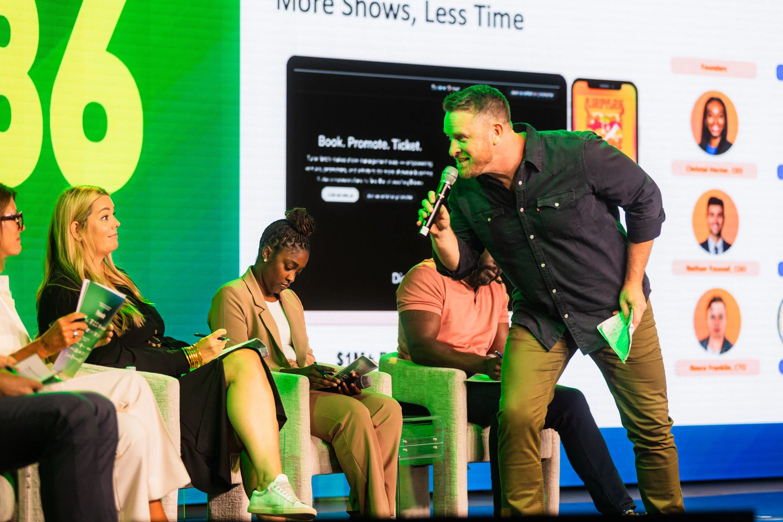 Chris McAdoo holding a microphone and speaking on a stage with a large screen behind him. Four people are seated on stage, three women and one man, looking at his display or listening attentively.