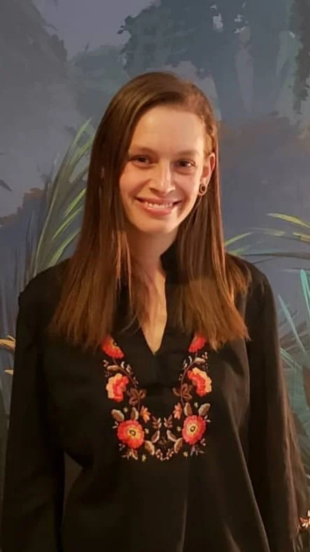 A woman with long straight brown hair, smiling, wearing a black embroidered top with red and pink floral designs, standing in front of a painted nature background.