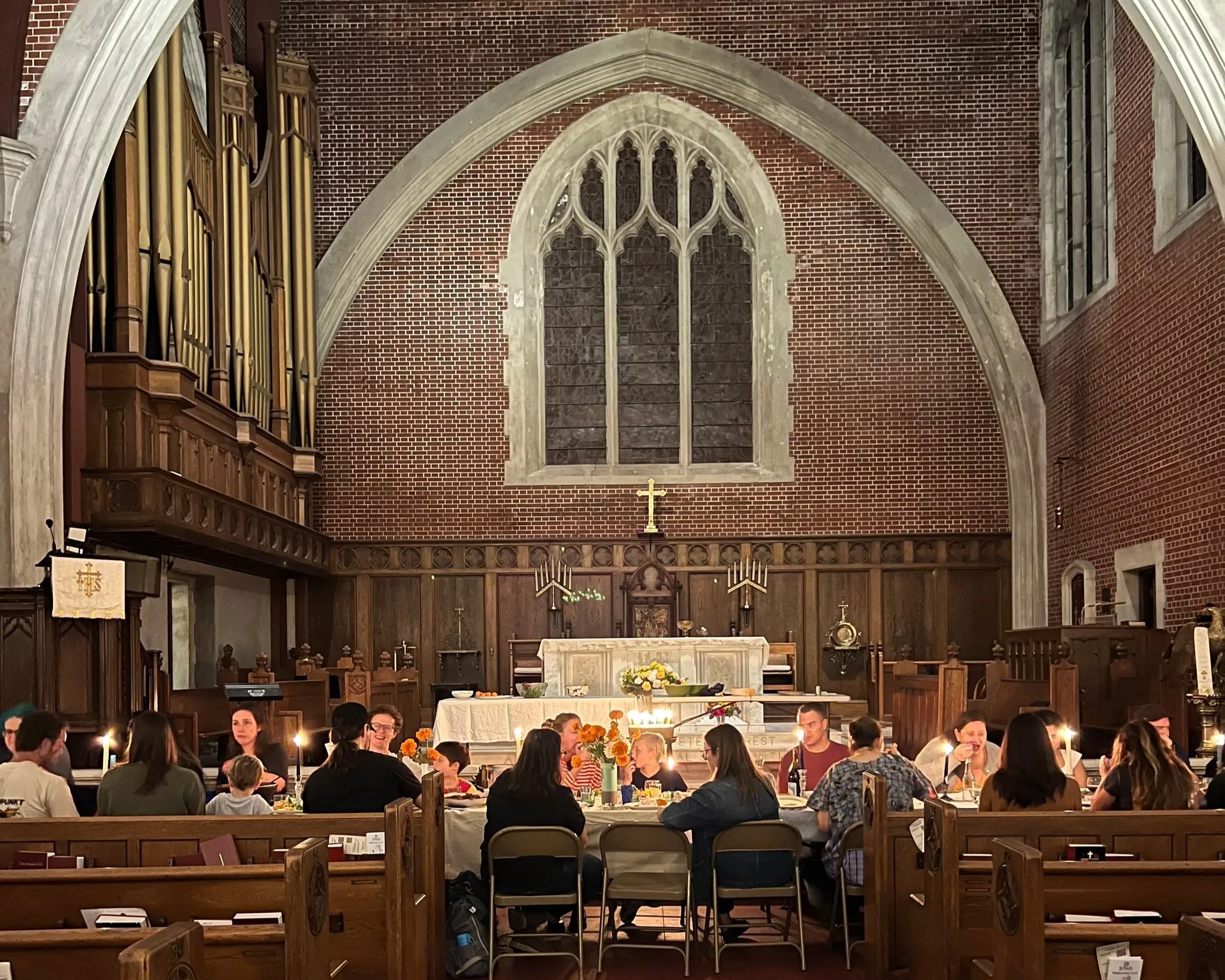 A church interior with a diverse group of people gathered around tables, some with lit candles, and an altar decorated with flowers. The background features a large stained glass window, brick walls, and wooden architectural details, including an organ and a crucifix.