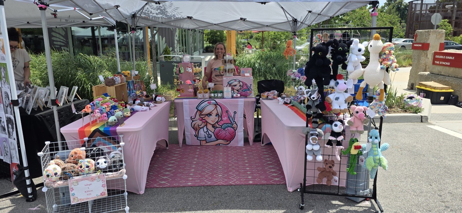 The Knotty Nurse Crochet booth at a craft show, with plush animals on pink-covered tables and Krista smiling behind the counter