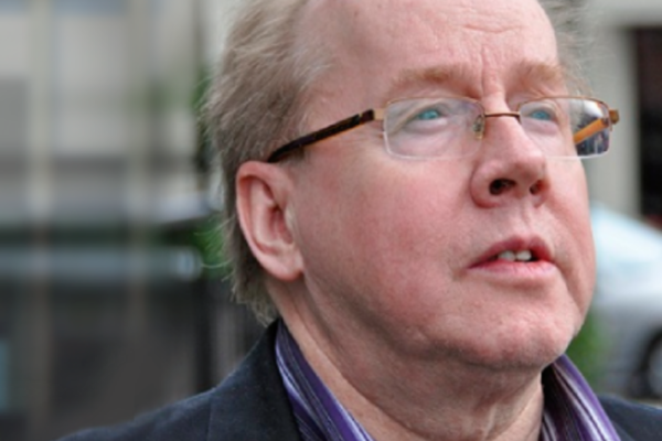 Close-up of a middle-aged man with glasses, light-colored hair, and wearing a dark jacket, outdoors in an urban setting.