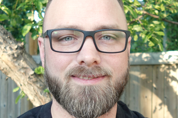 Close-up of a man with glasses and a beard smiling outdoors with green foliage and a wooden fence in the background.