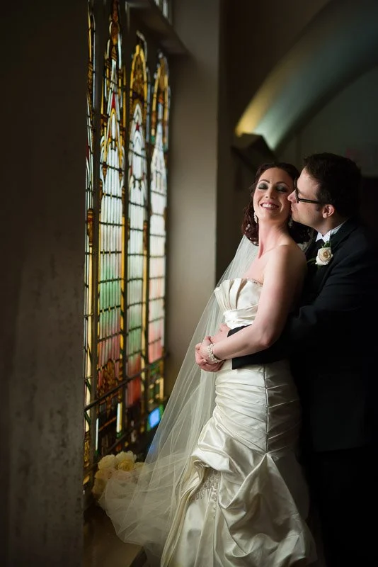 A bride and groom stand together near stained glass windows in a church, smiling and sharing a tender moment on their wedding day.