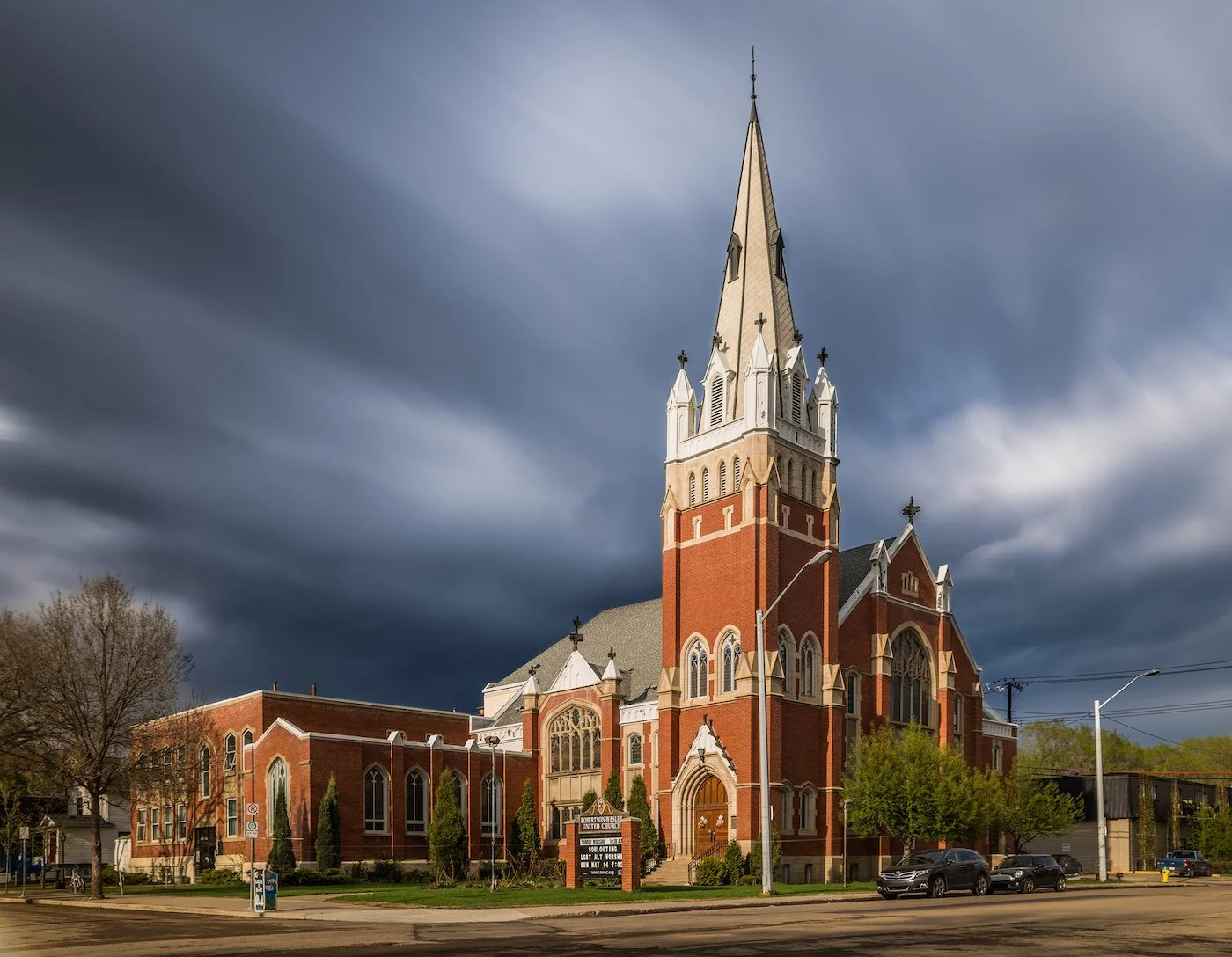 A large brick church with a tall steeple and Gothic-style windows under dark, stormy skies, surrounded by trees and parked cars.