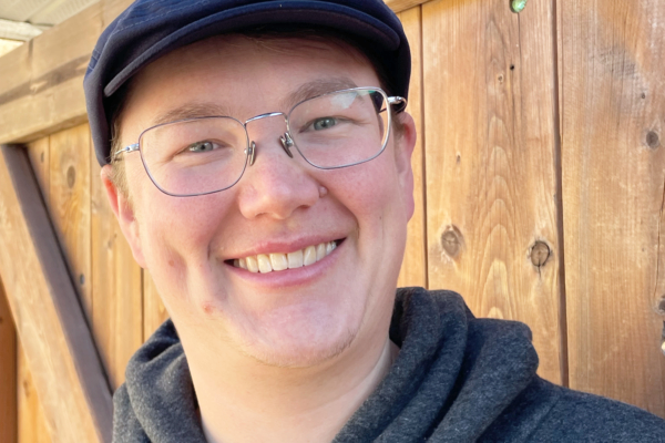Young man smiling wearing glasses and a cap, standing outdoors in front of a wooden fence.