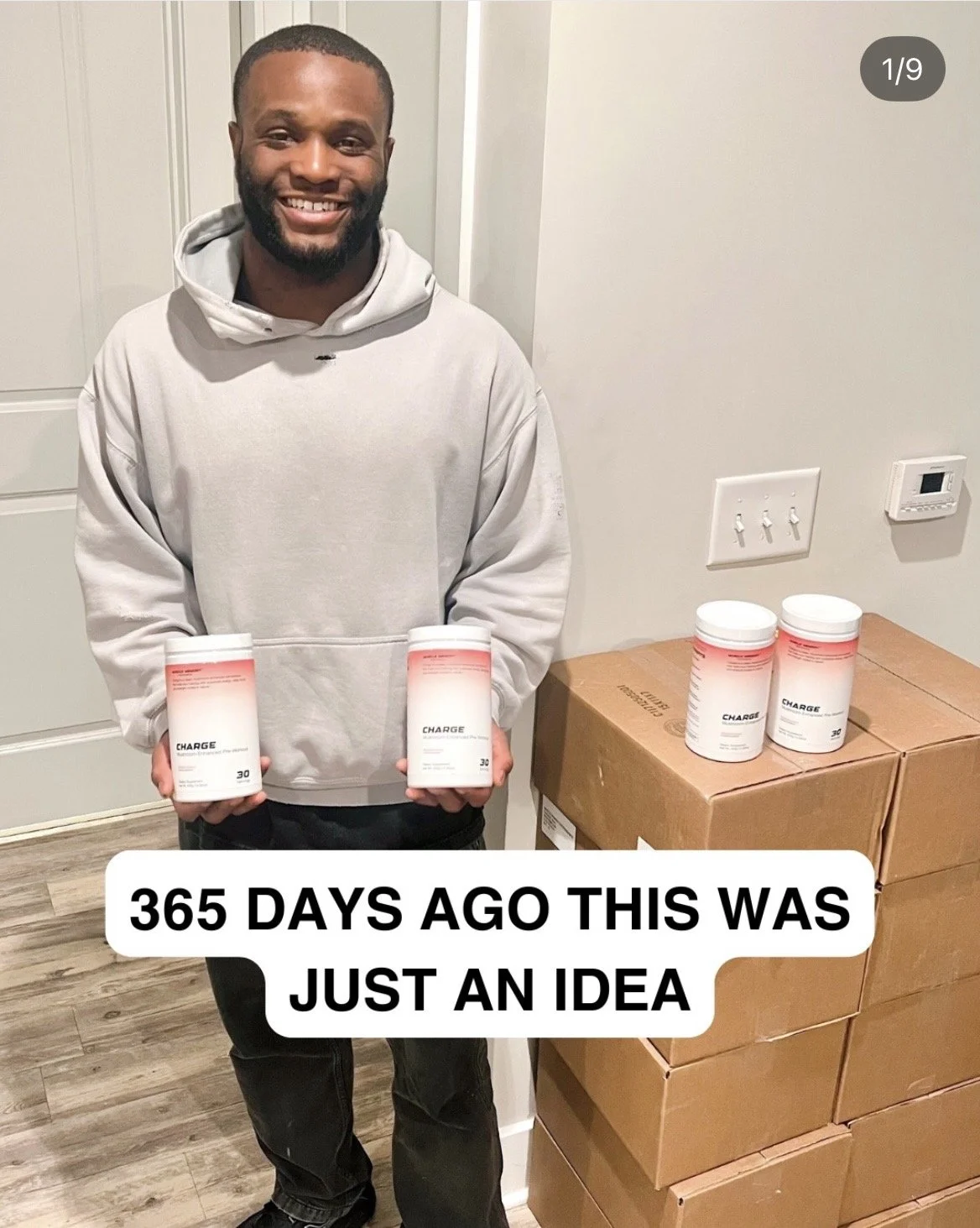 Ralph Patton III smiling and holding two tubs of CHARGE pre-workout in front of stacked shipping boxes of inventory