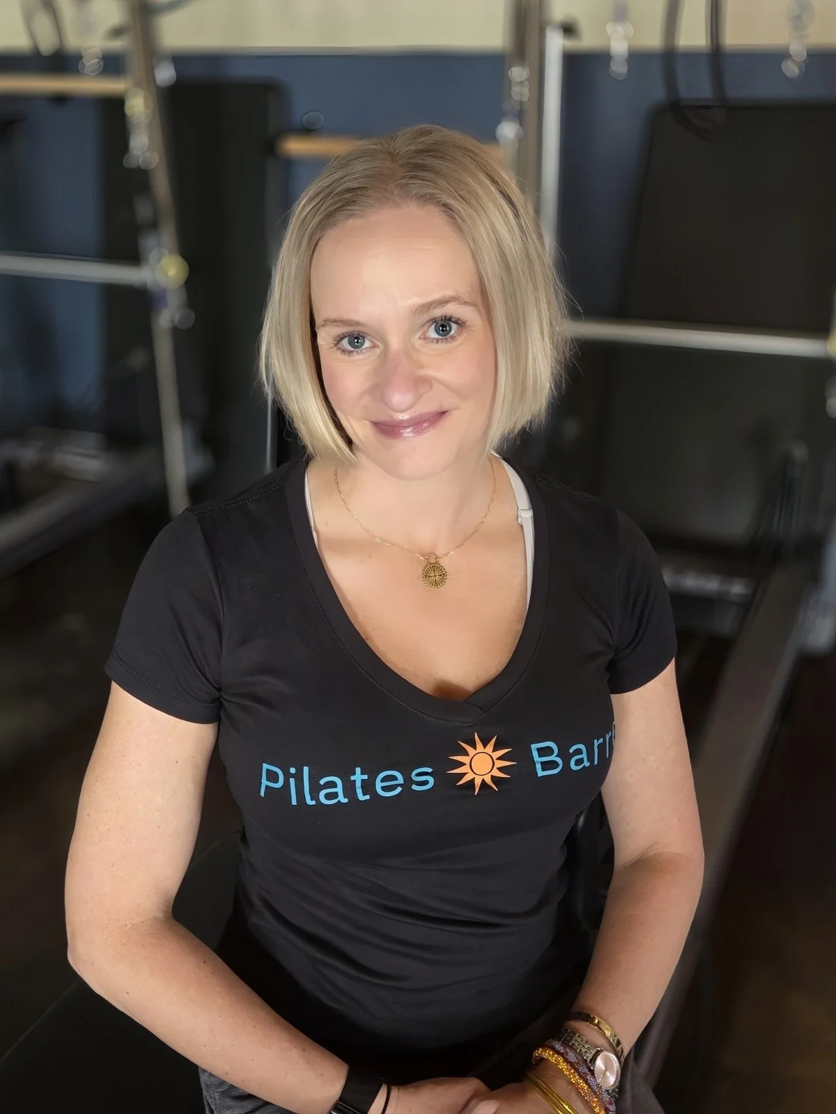 A woman with blonde hair, wearing a black T-shirt with 'Pilates' and 'Barre' written on it, sitting indoors with resistance bands and chairs in the background.