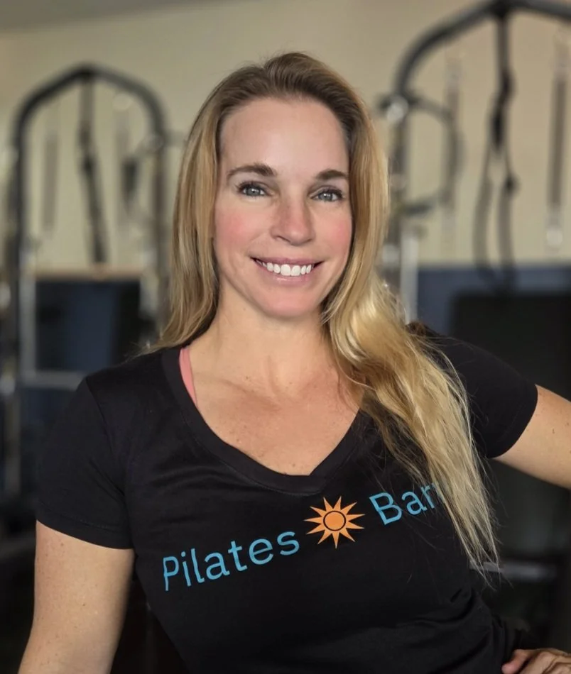 A smiling woman with long blonde hair in a Pilates studio, wearing a black T-shirt that says 'Pilates Bar' with a sun logo.