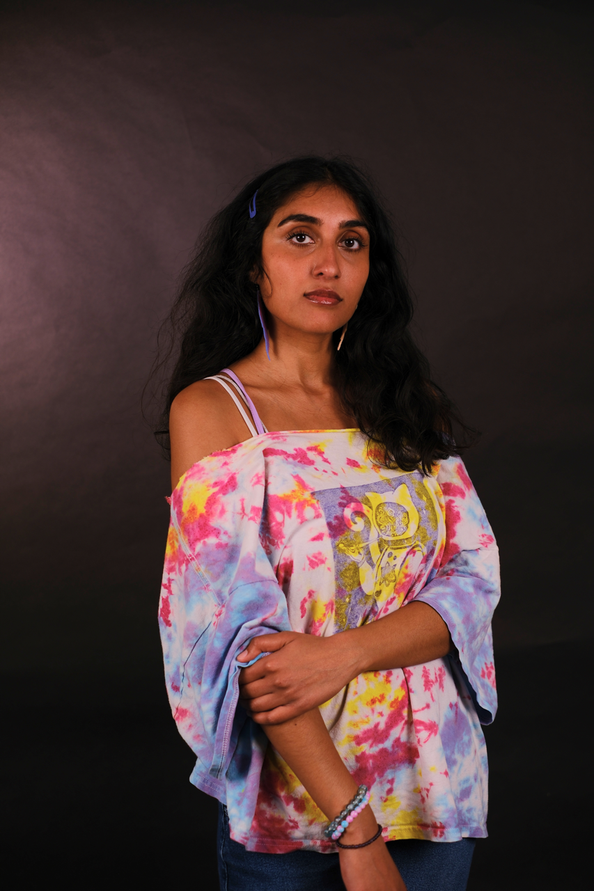A young woman with long dark hair, wearing a tie-dye off-shoulder top with a print on the front, against a dark background.