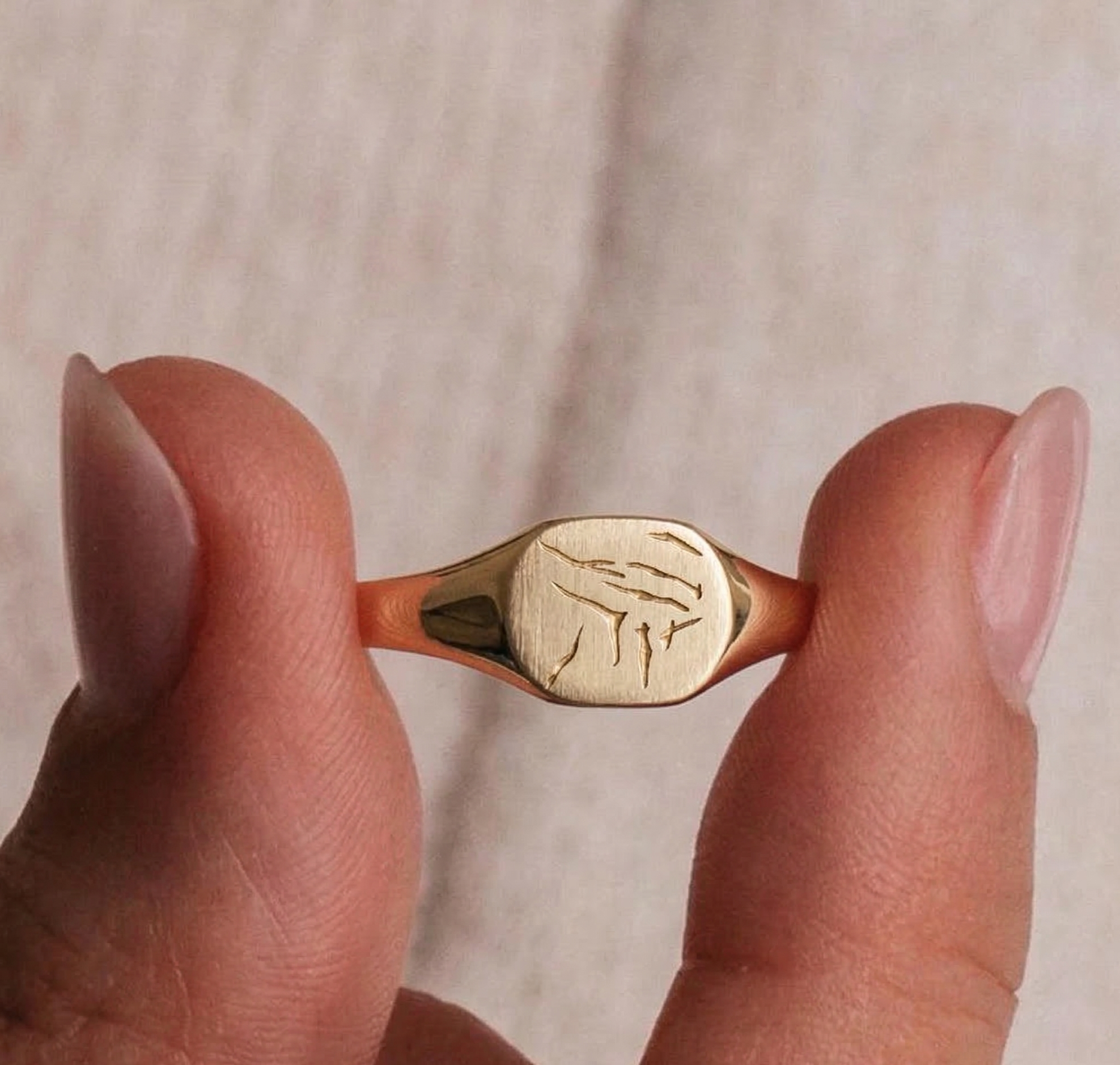 Close-up of a person holding a gold ring with engraved lion face design between thumb and index finger against a neutral background.